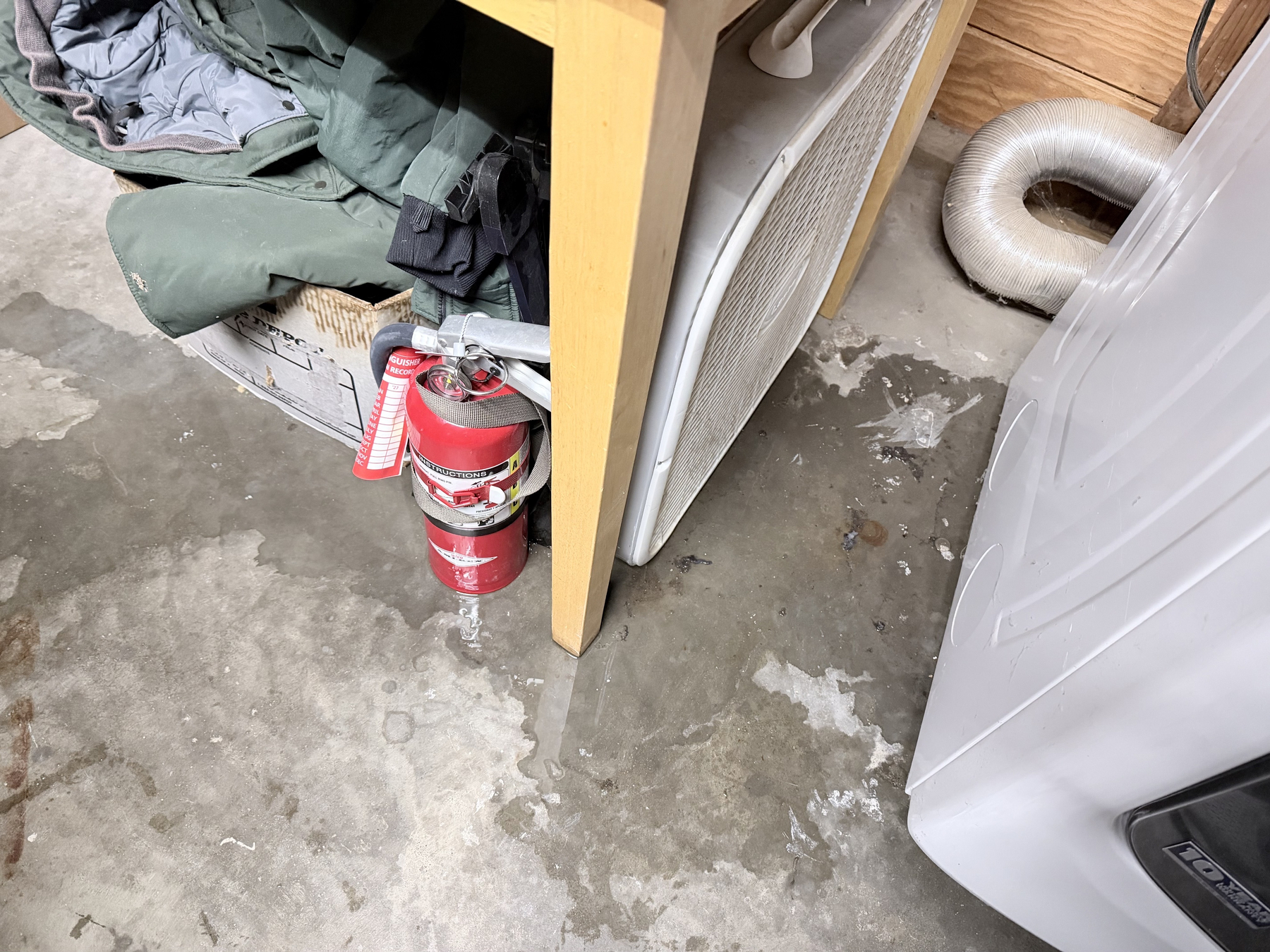 IMG_0565.jpeg An unfinished garage with a small leak on the floor under and beside a dryer. The side of a dryer. then some clear space, then a table with box fan, fire extinguisher, some old coats under it. The water ran downhill the concrete pad to the left.