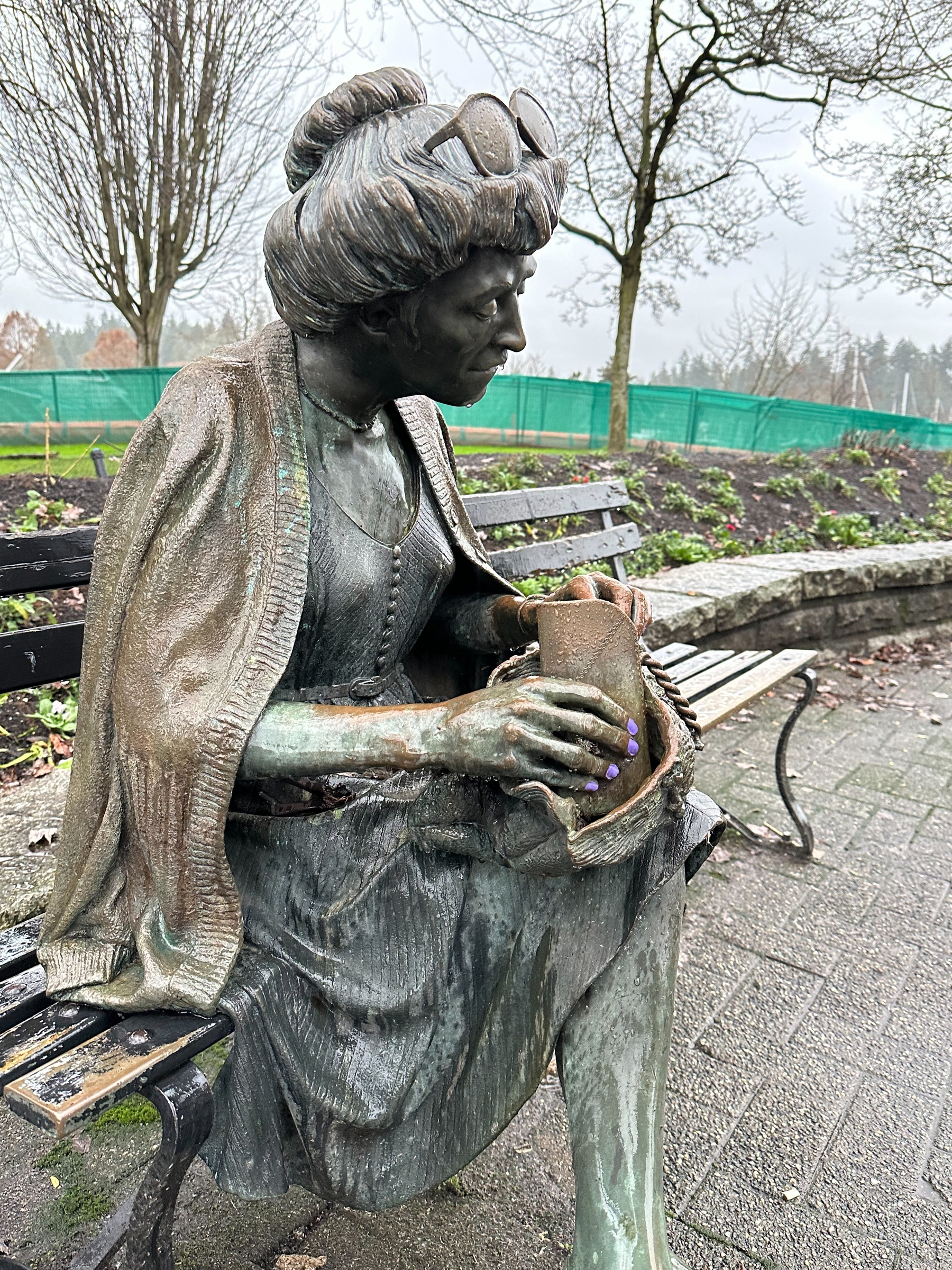 At Stanley Park, Vancouver, with copper sculpture of modestly dressed, cross legged, senior lady, with bunned hair and big sun glasses, that has had her nails painted violet and looks like she's crying because of the rain drops