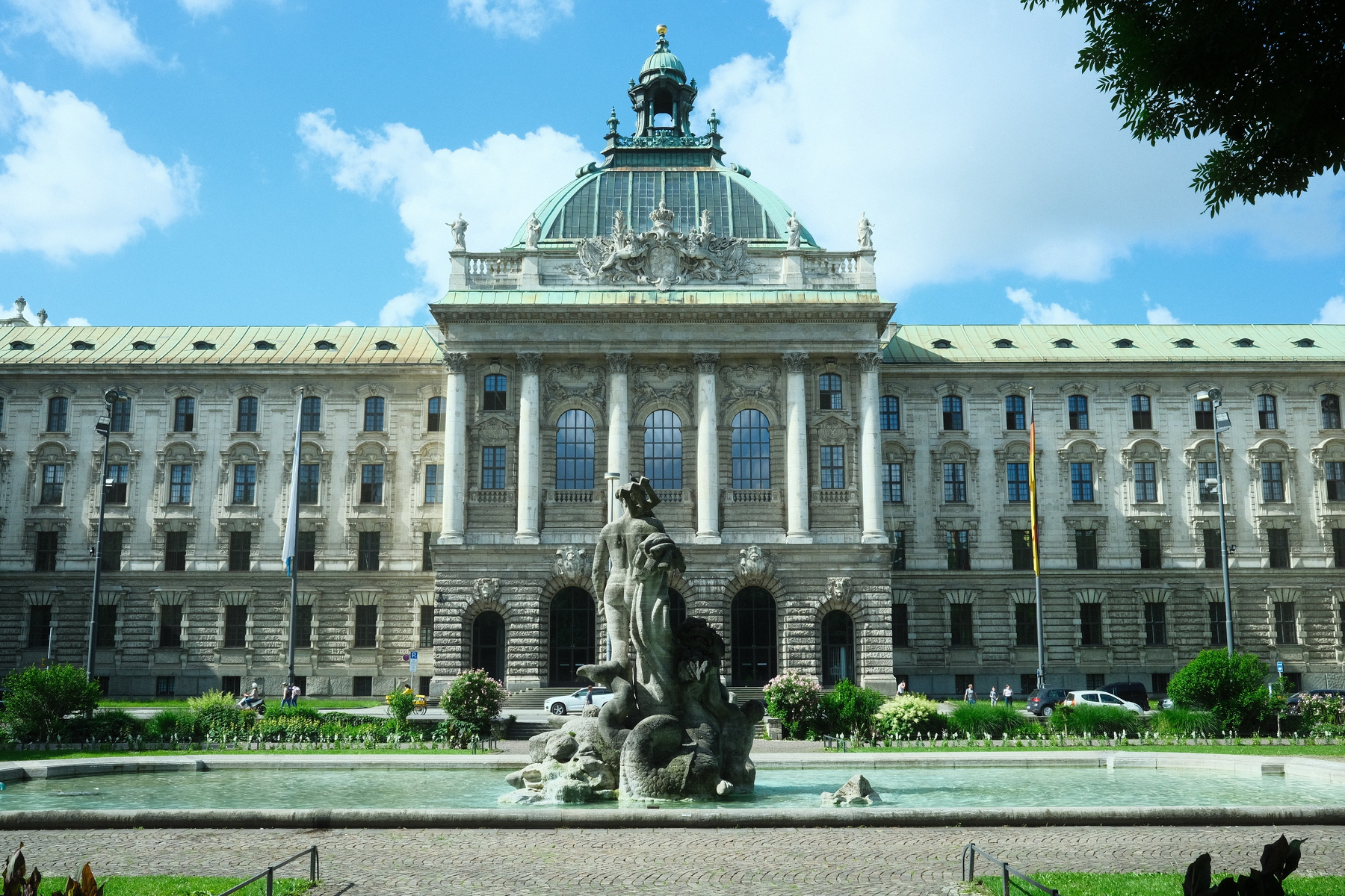 The rear of the Neptune Fountain, with Neptune's rear end prominent in front of the Palace of Justice. It's a nice blue but cloudy day, and the sky with clouds reflect in the palace's oversized windows. The copper and glass dome reaches toward the sky.