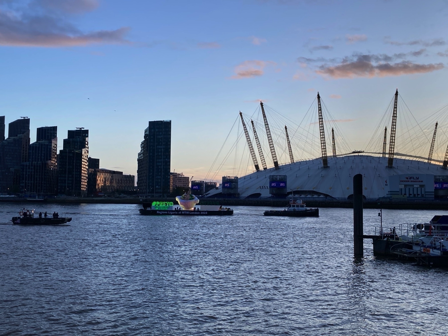 A float advertising pesto is being pulled up the Thames by a tugboat past the O2 dome on the Greenwich Peninsula as the sun sets.