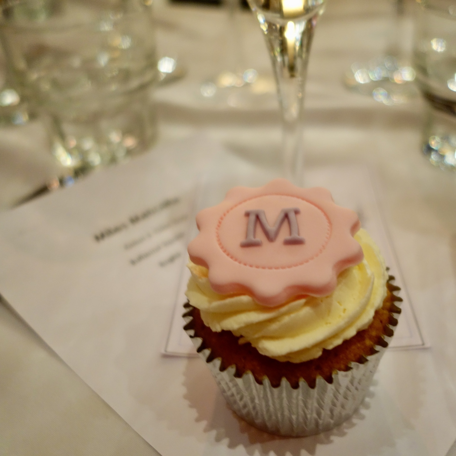 A cupcake with the initial M on top of a menu, surrounded by wine glasses, offered as a birthday favour