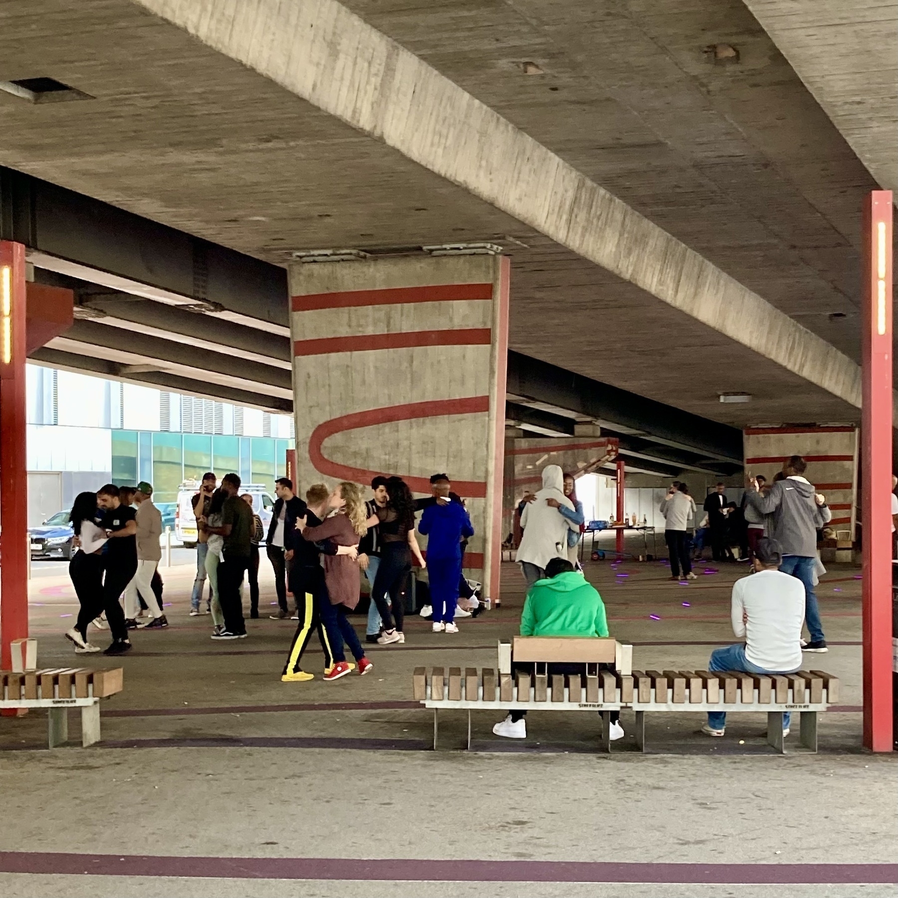 Couples dance together under a concrete flyover, decorated with red swooshes, people sitting on benches look on. Terry Spinks Place, Canning Town, East London