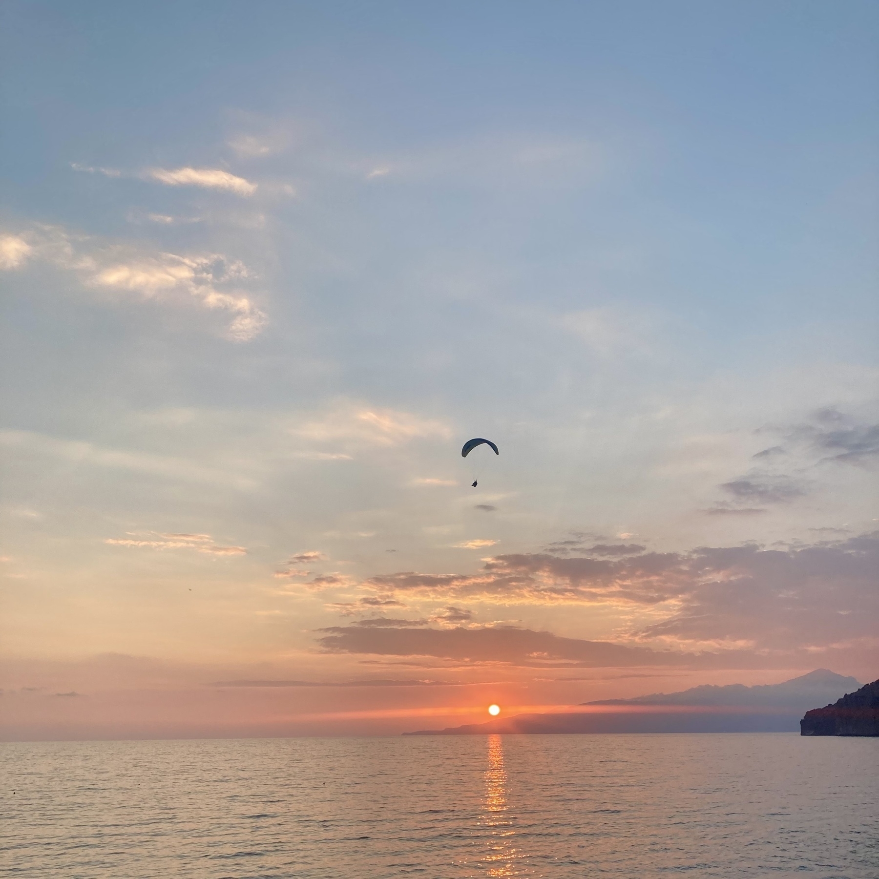 A paraglider descends towards the beach over the sea, with the sun setting behind the shoulder of a mountain in the background
