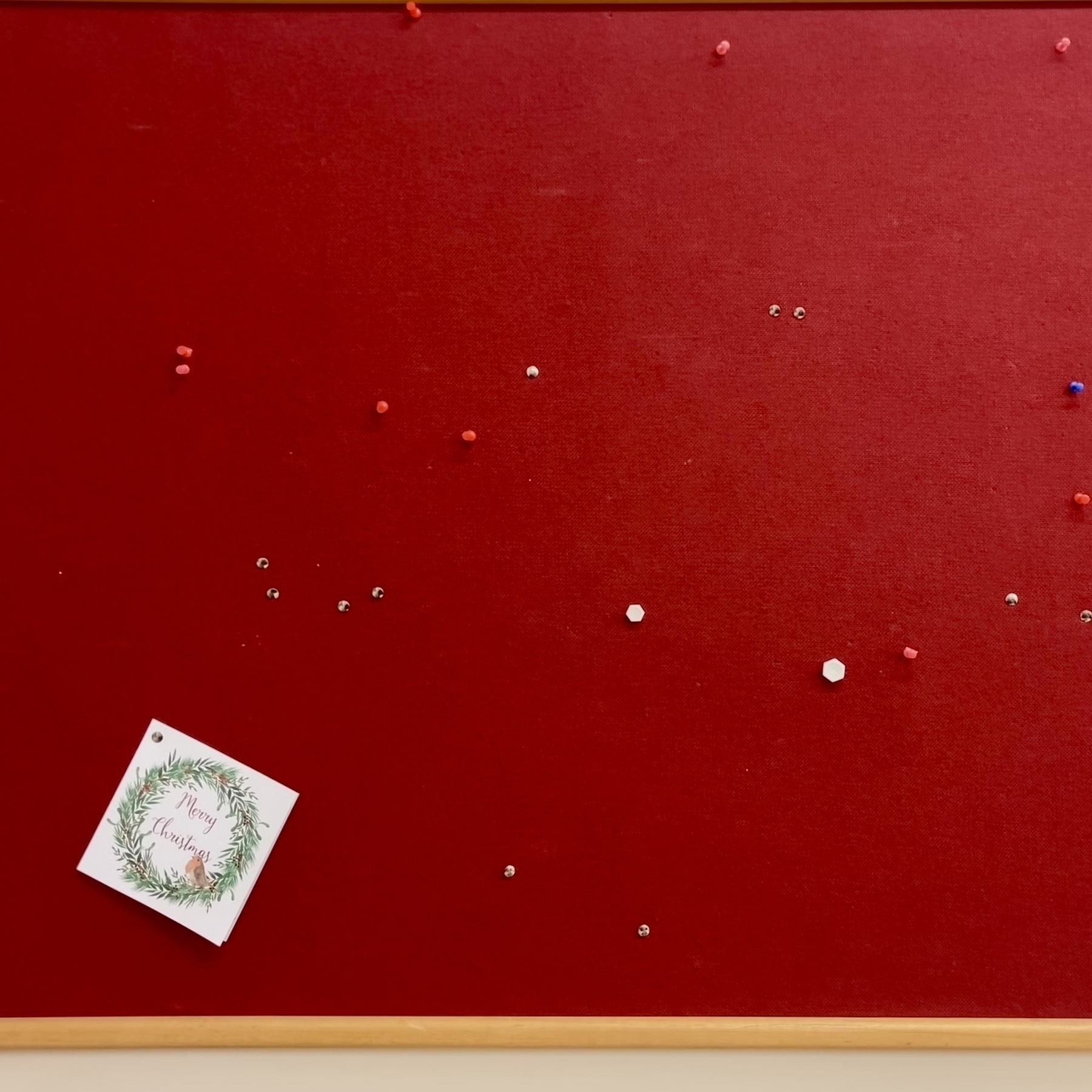 A solitary, forgotten Christmas card on a Christmas-red noticeboard stuck with pins