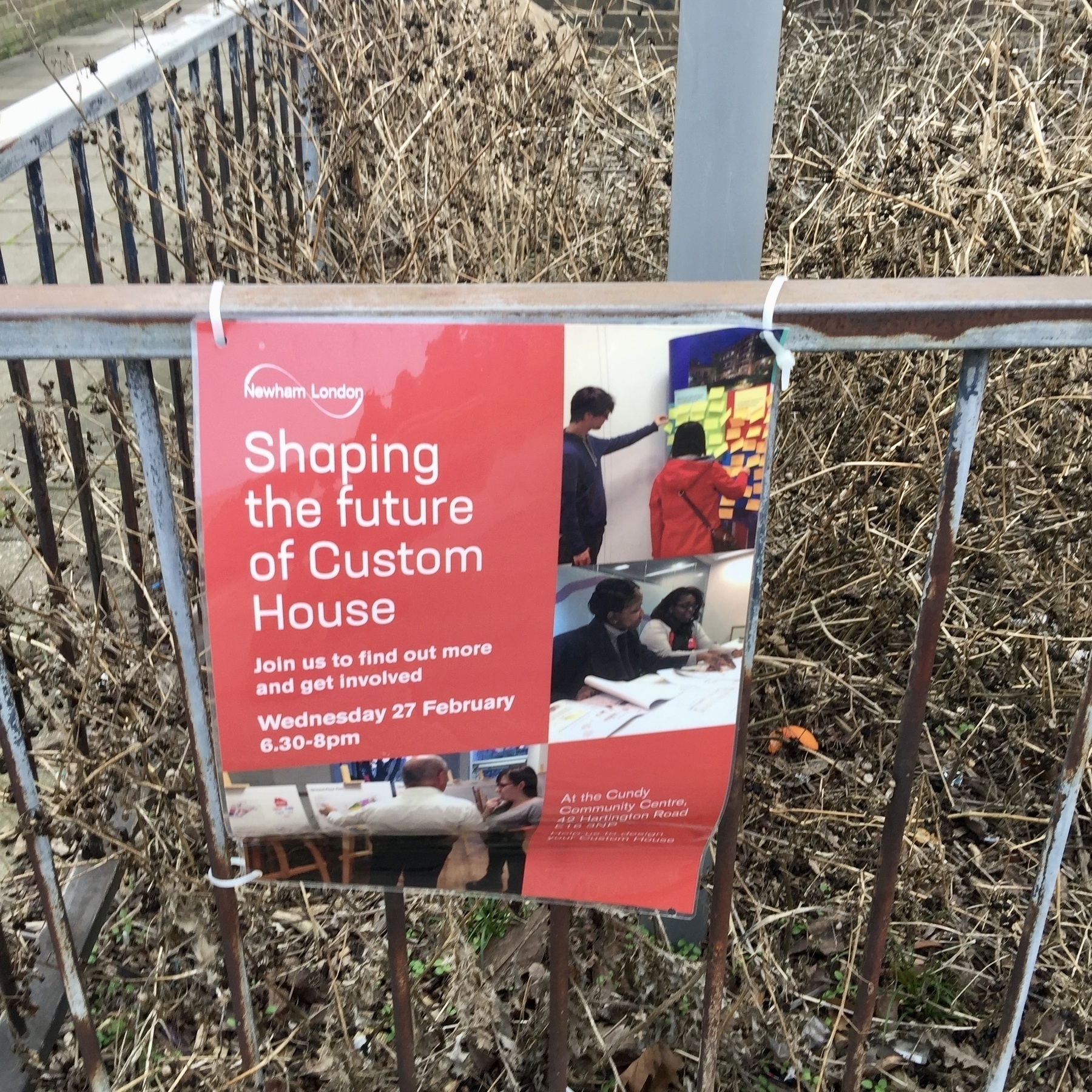 A red poster advertising a community event fixed to a dilapidated metal fence, behind which is dessicated overgrown grass and weeds.