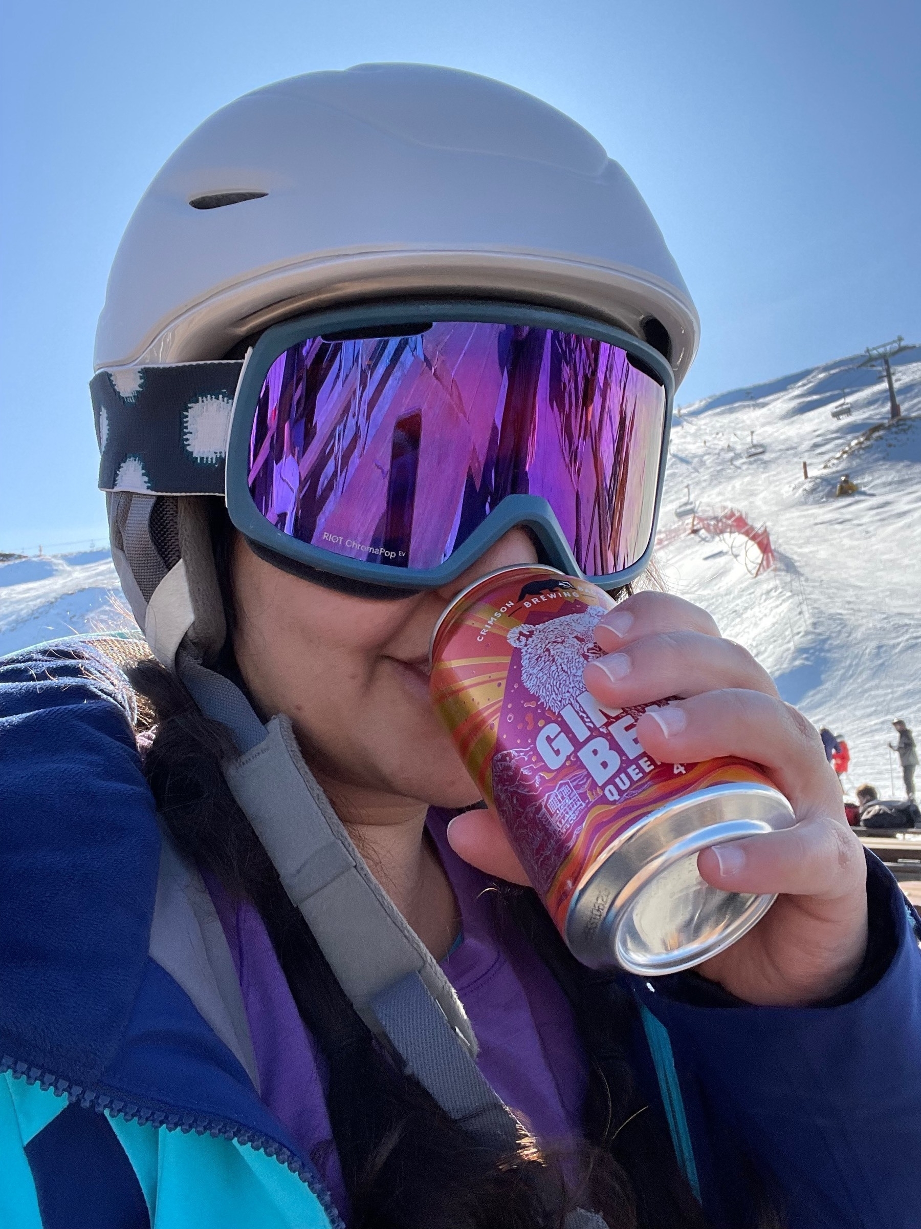 Meera in a white ski helmet and reflective purple ski goggles in front of a snow-covered mountain drinking a Ginger Bear from an orange and maroon can.