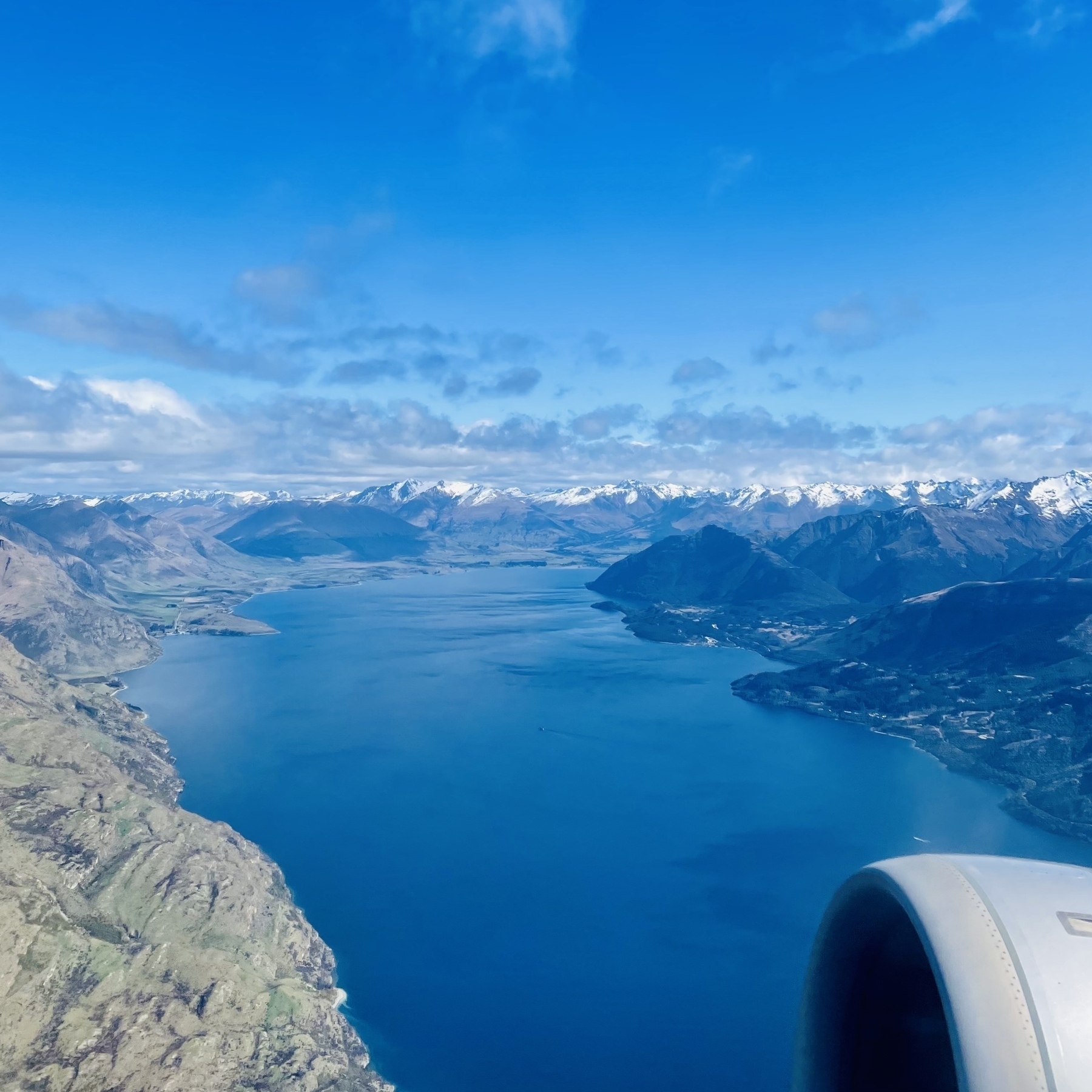 A New Zealand fjord. Blue water in the middle, green mountains on both sides, a full horizon of snow-capped peaks in the distance, and a plane engine in the lower-right.