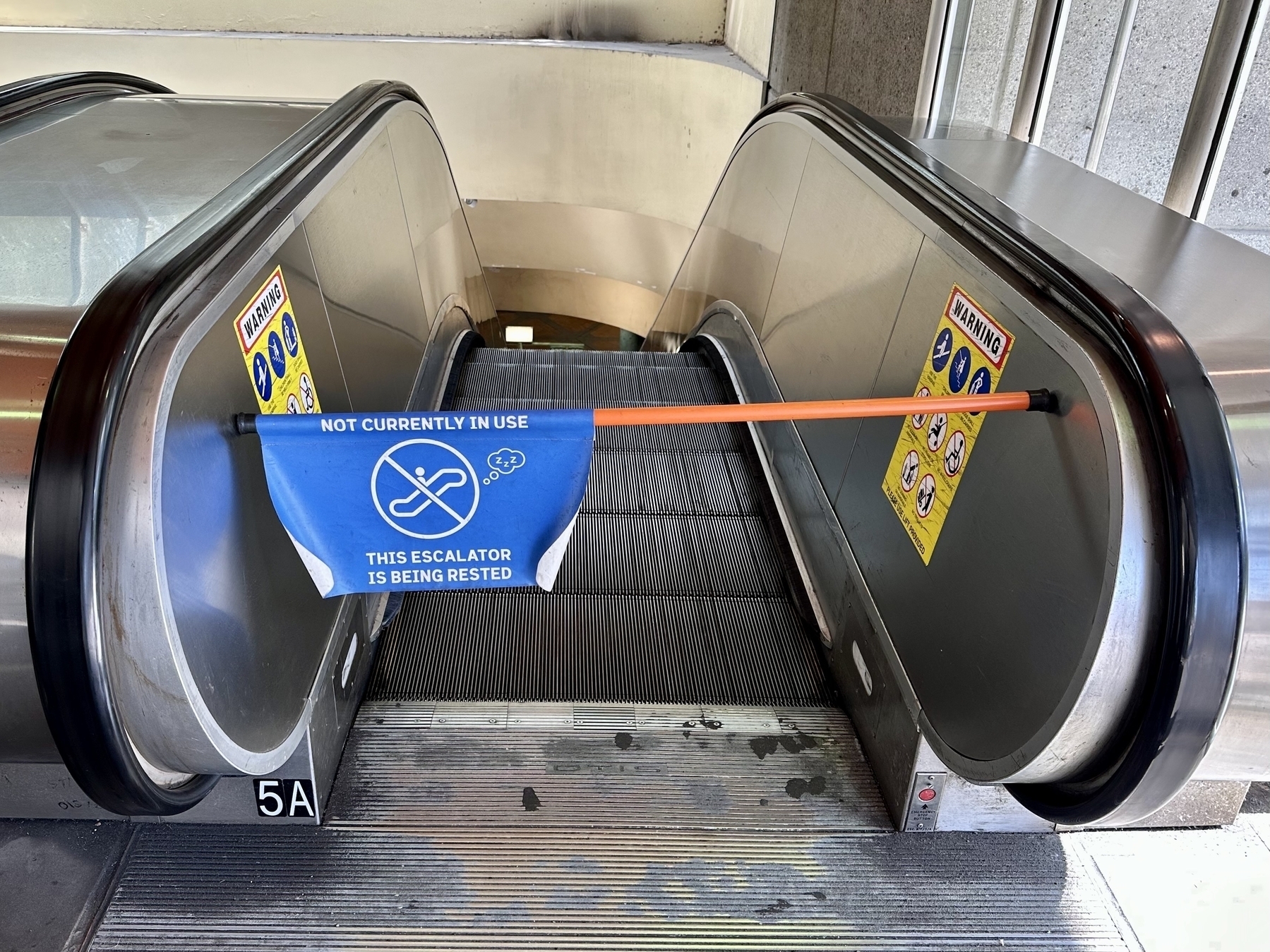 Top of an escalator blocked by an orange bar with a blue flag reading in white letters, “Not currently in use, this escalator is being rested.” The flag also has an escalator icon with a thought bubble filled with Z’s.