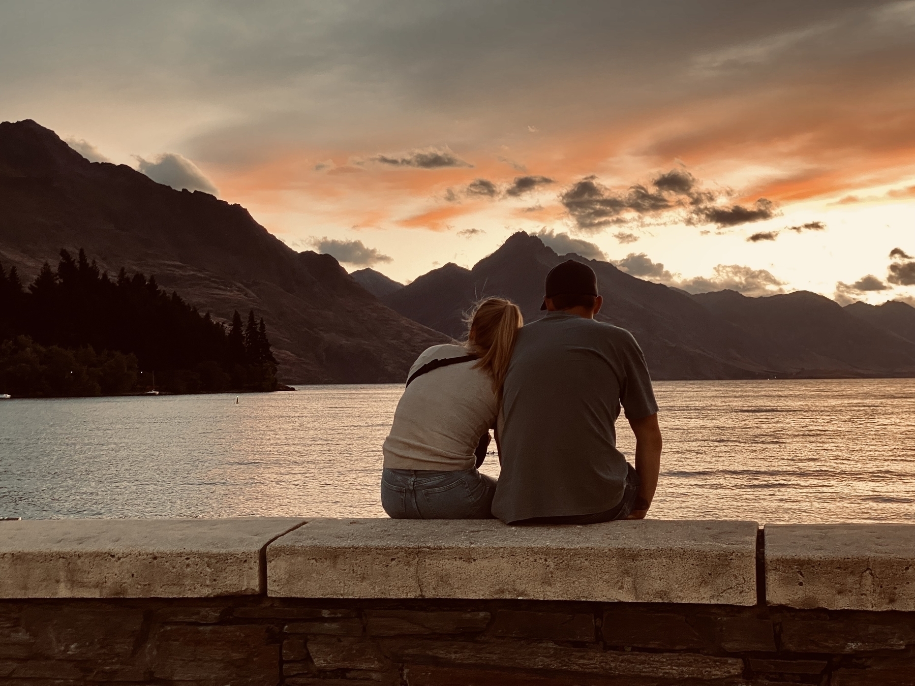 A young woman rests her head on a young man’s shoulder as they watch the sunset over Lake Wakatipu.