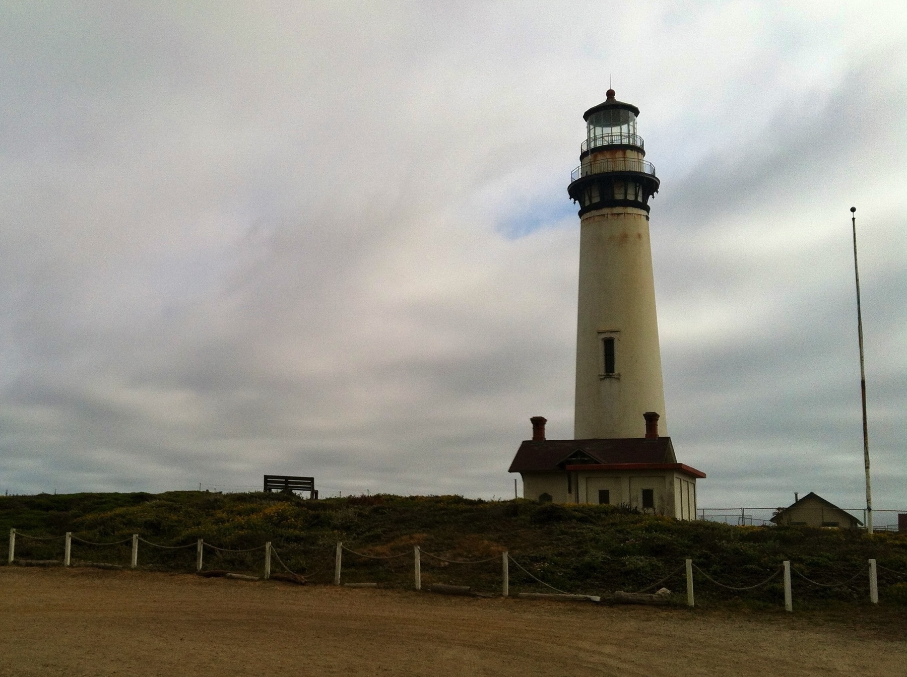 A traditional white lighthouse, faded and spotted with rust, atop a house with a gabled roof and two chimneys. An empty bench looks out onto the ocean, while an empty sand parking lot and overcast sky complete the gloomy picture.