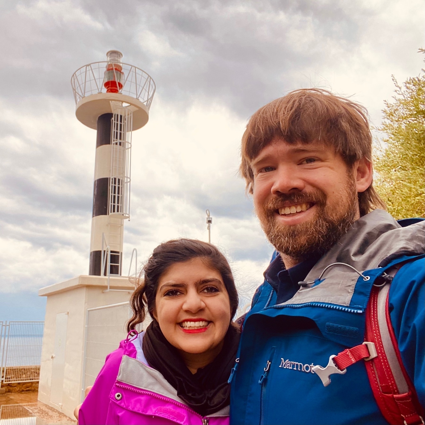 Photo of me and Meera in front of Far de la Mola lighthouse, a thin tower on a cliff with wide horizontal bands of black and white and a bright red top.