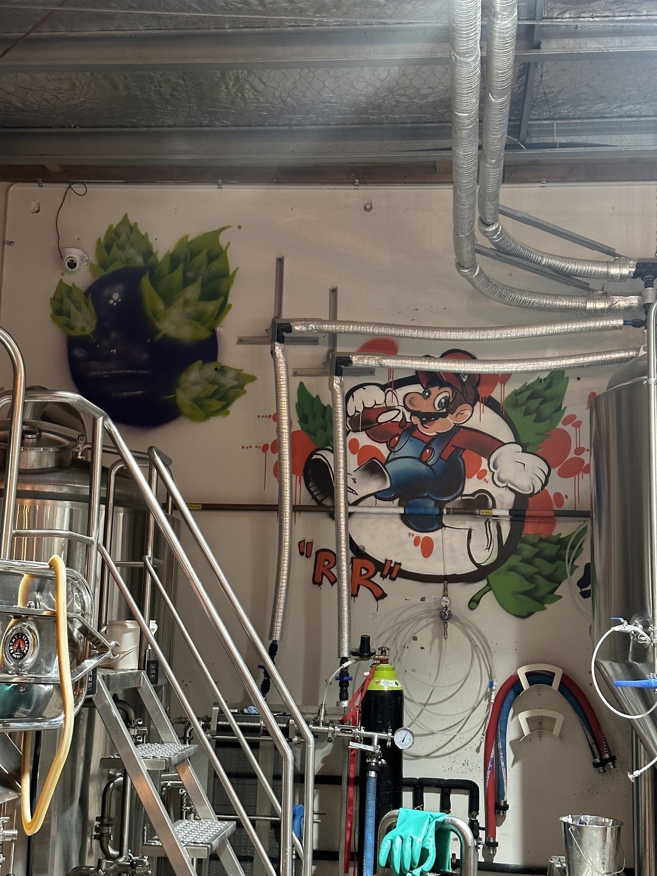 Two wall murals behind brewery vats. The left, a black planet orbited by giant hops. The right, Mario bursting through the wall, surrounded by hops, with the letters RxR in the bottom-left.