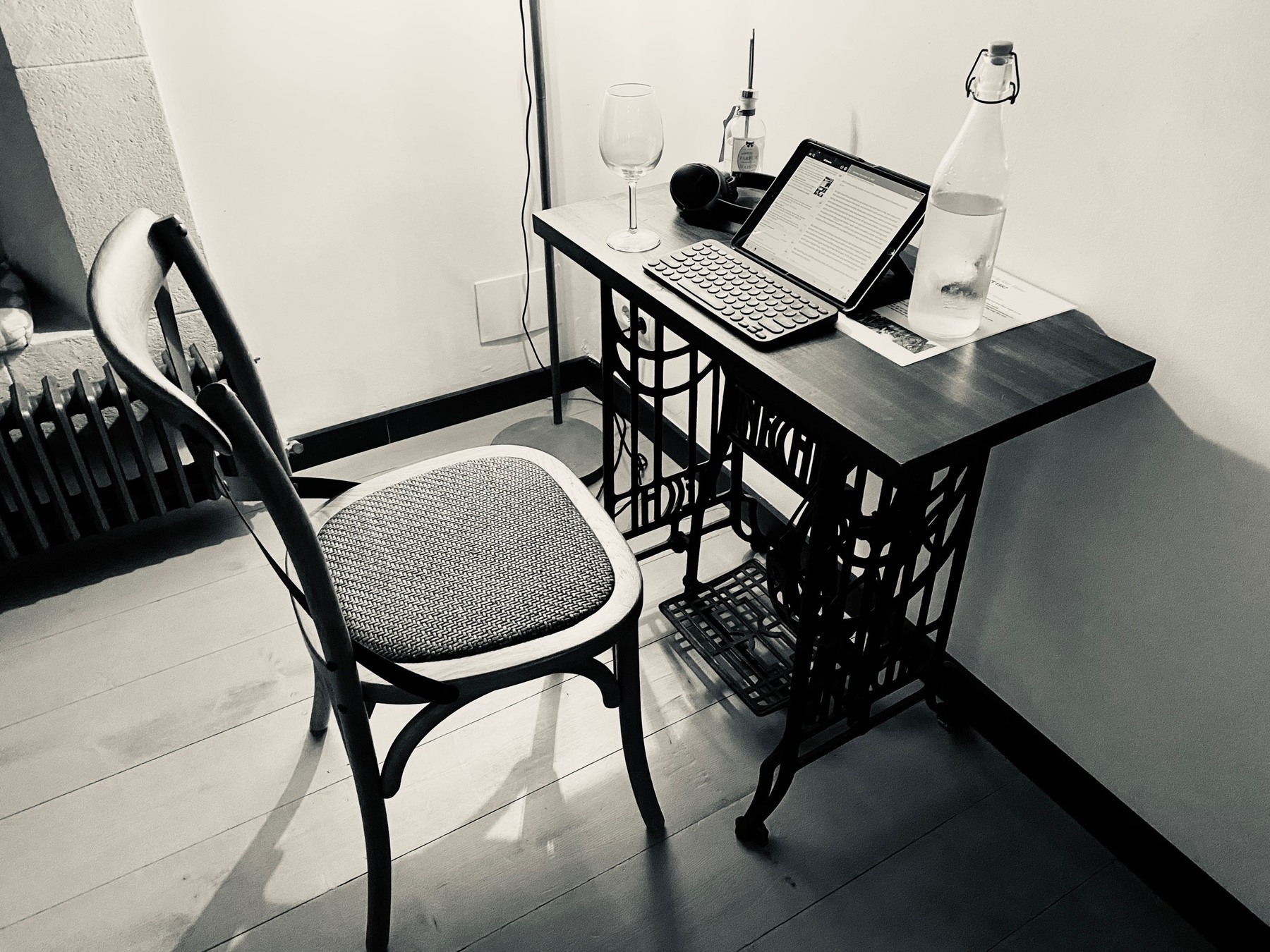Black-and-white photo of a wooden writing desk made of an old sewing machine, topped by an iPad, keyboard, water bottle, and wine glass.