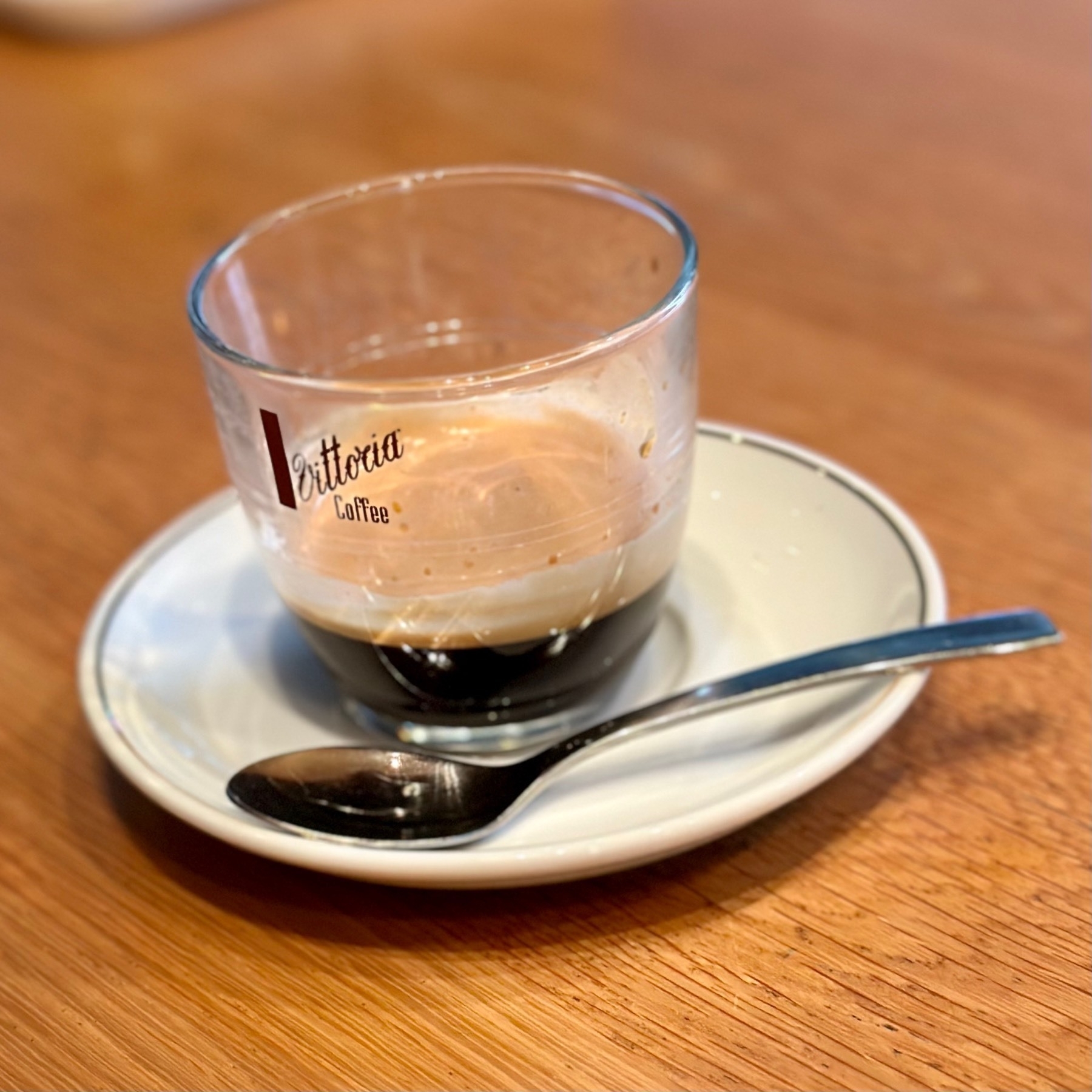A double espresso shot in a too-large Vittoria coffee tasse, on a saucer with a spoon.