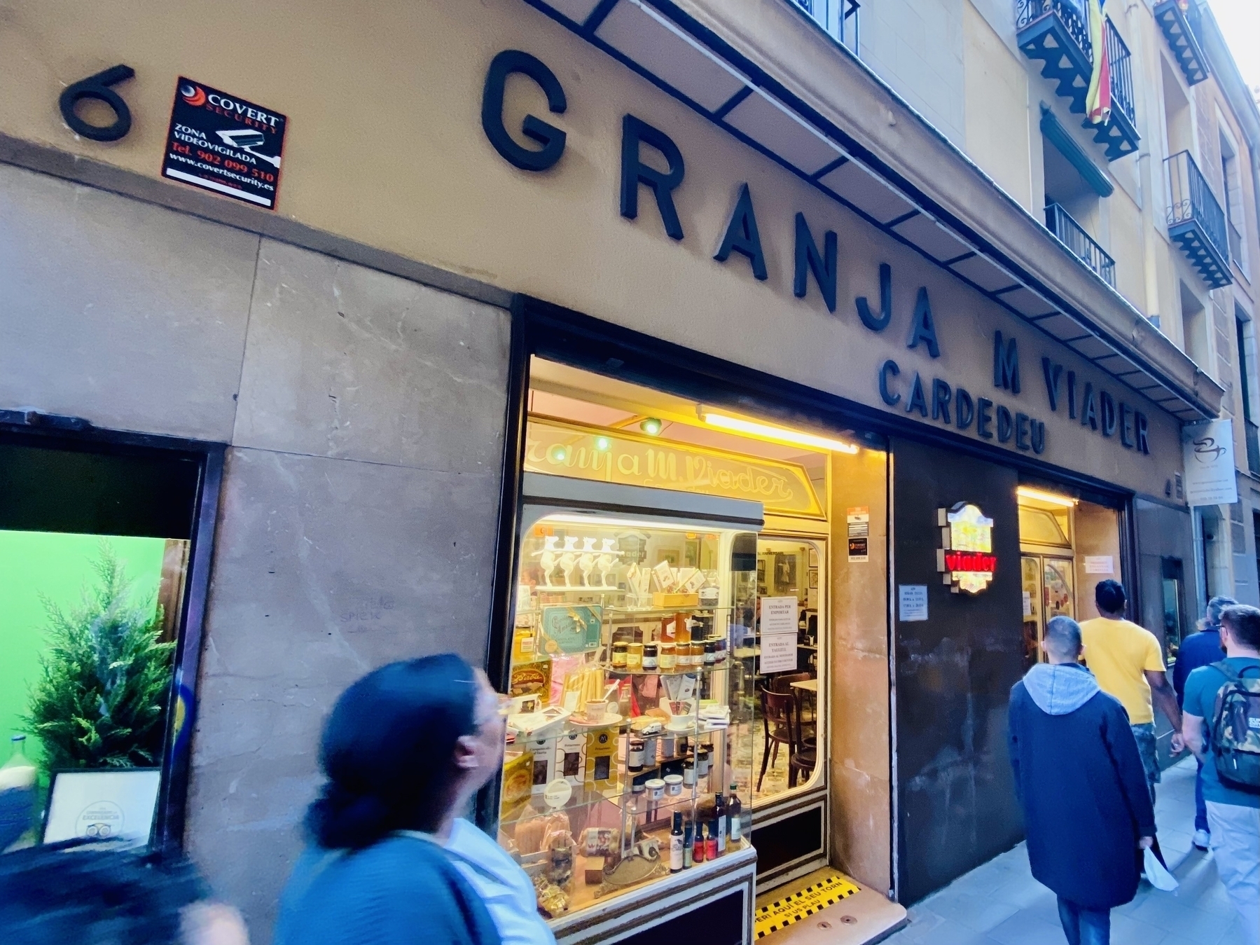 A few people walk past the outside of Granja M. Viader. A window display shows chocolates, jams, and wines for sale.
