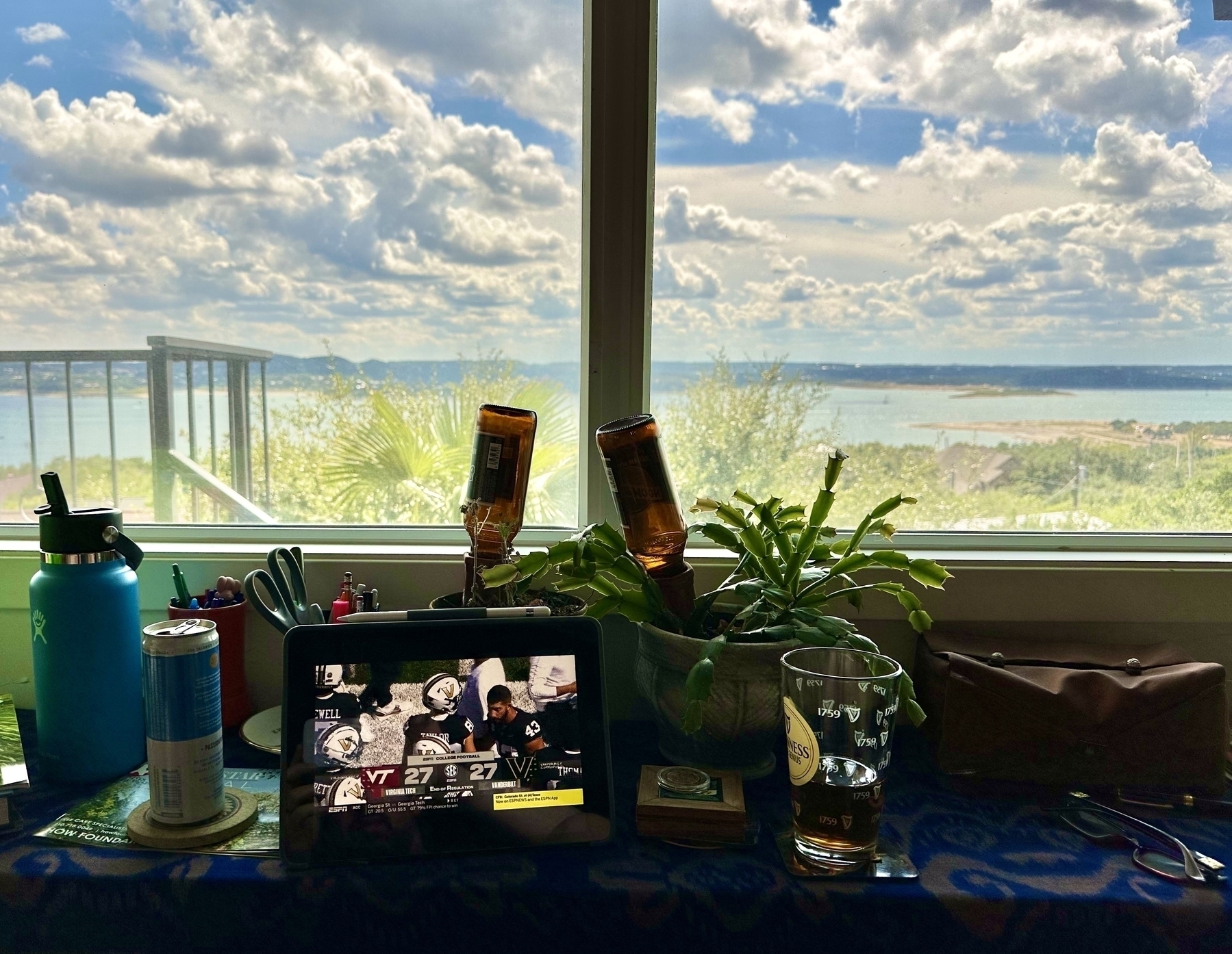 An iPad showing VT at Vanderbilt on a table in front of a window looking out onto a lake with bright blue skies and clouds. Seltzer on one side, beer in a pint glass on the other. Score is 27-27, sorry Hokies.