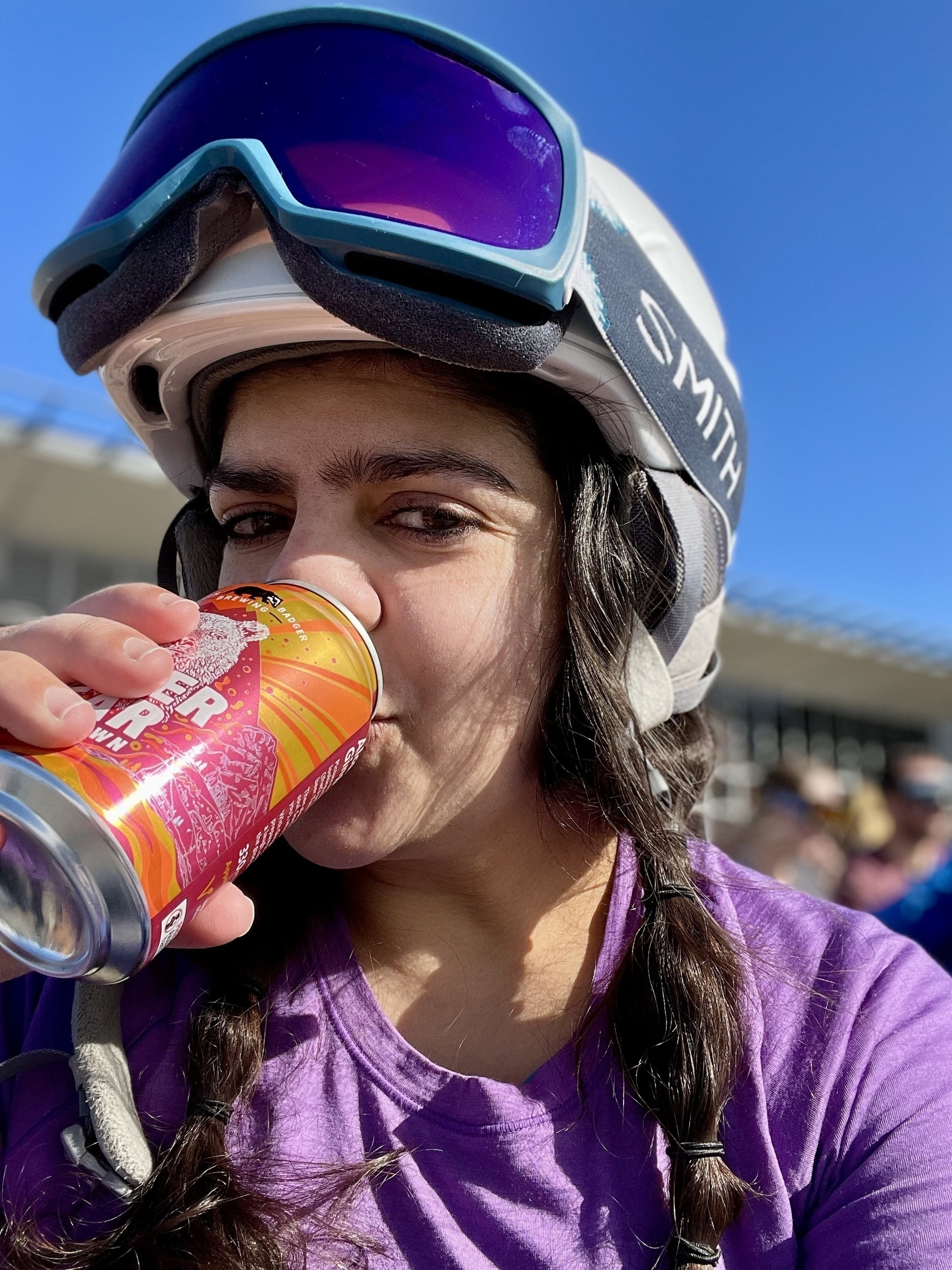 Meera in a ski helmet and goggles drinking a beer.