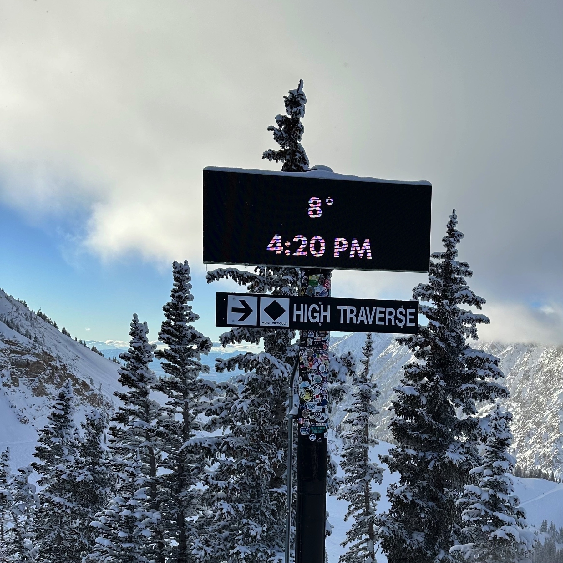 Digital sign reading 8°, 4:20PM. Black diamond trail sign for High Traverse. In the background, snow on fir trees in front of a mountain valley colored by the setting sun but topped with gray clouds.