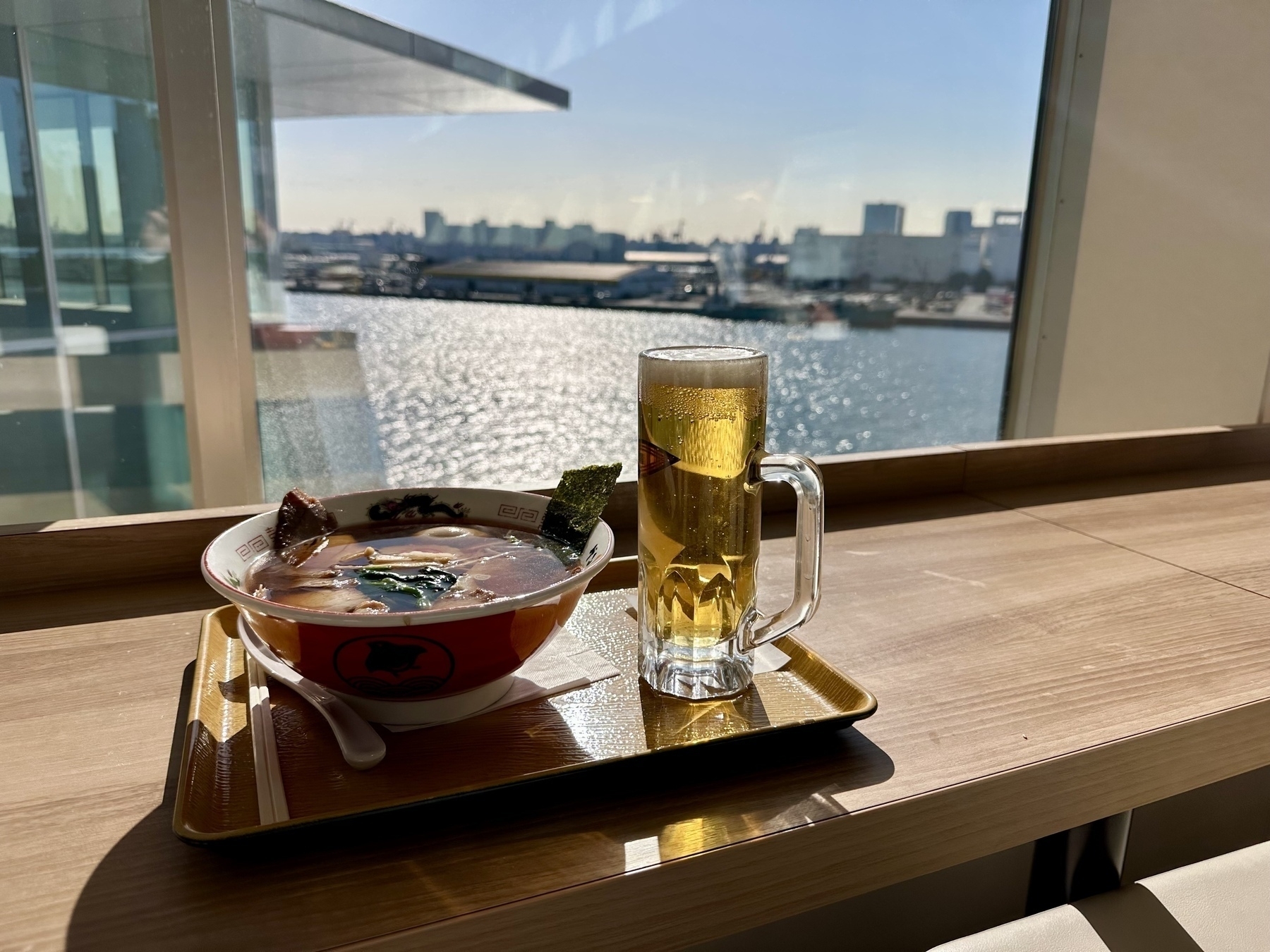 A bowl of ramen and a small stein of beer on a tray on a bar in front of a window looking out onto a port on Tokyo Bay.