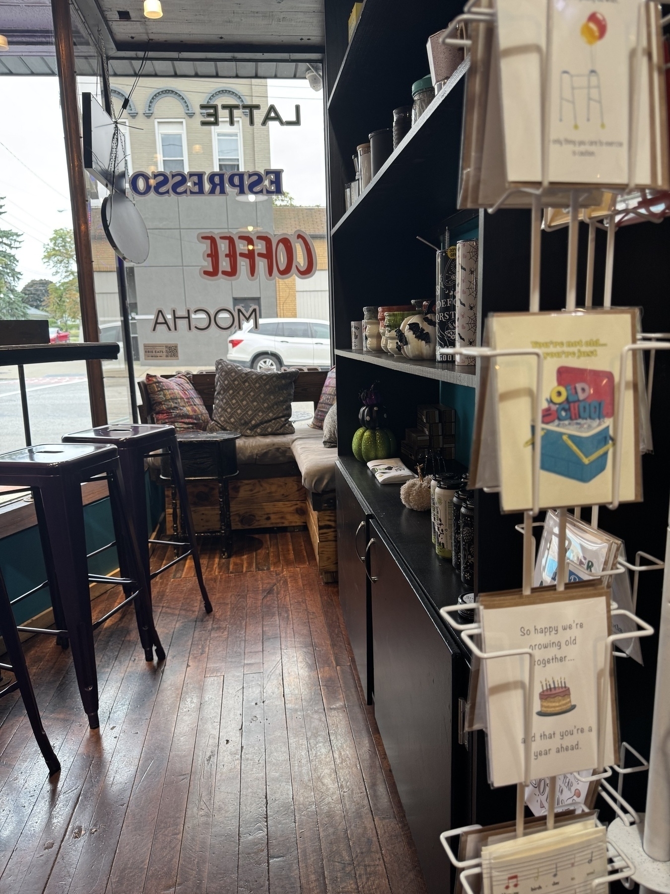 A cozy coffee shop interior with a seating area by the window and a display of greeting cards on the right. There’s a hutch on the right with mugs and coffee gear. The Word Coffee is painted on the window and a white car parked outside.