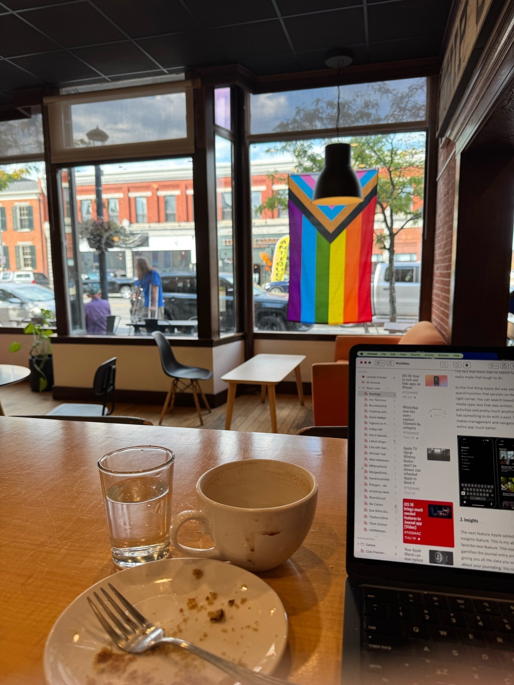 Coffee and MacBook Air on table with a view outside. The windows has a pride flag hanging on it&10;