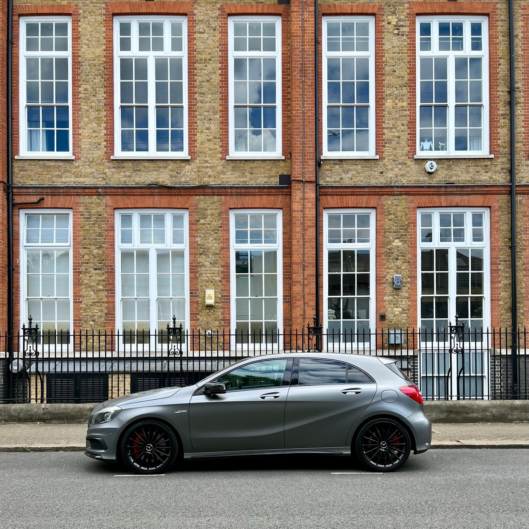 A gray car is parked on the street in front of a building with red brickwork and large white-framed windows.