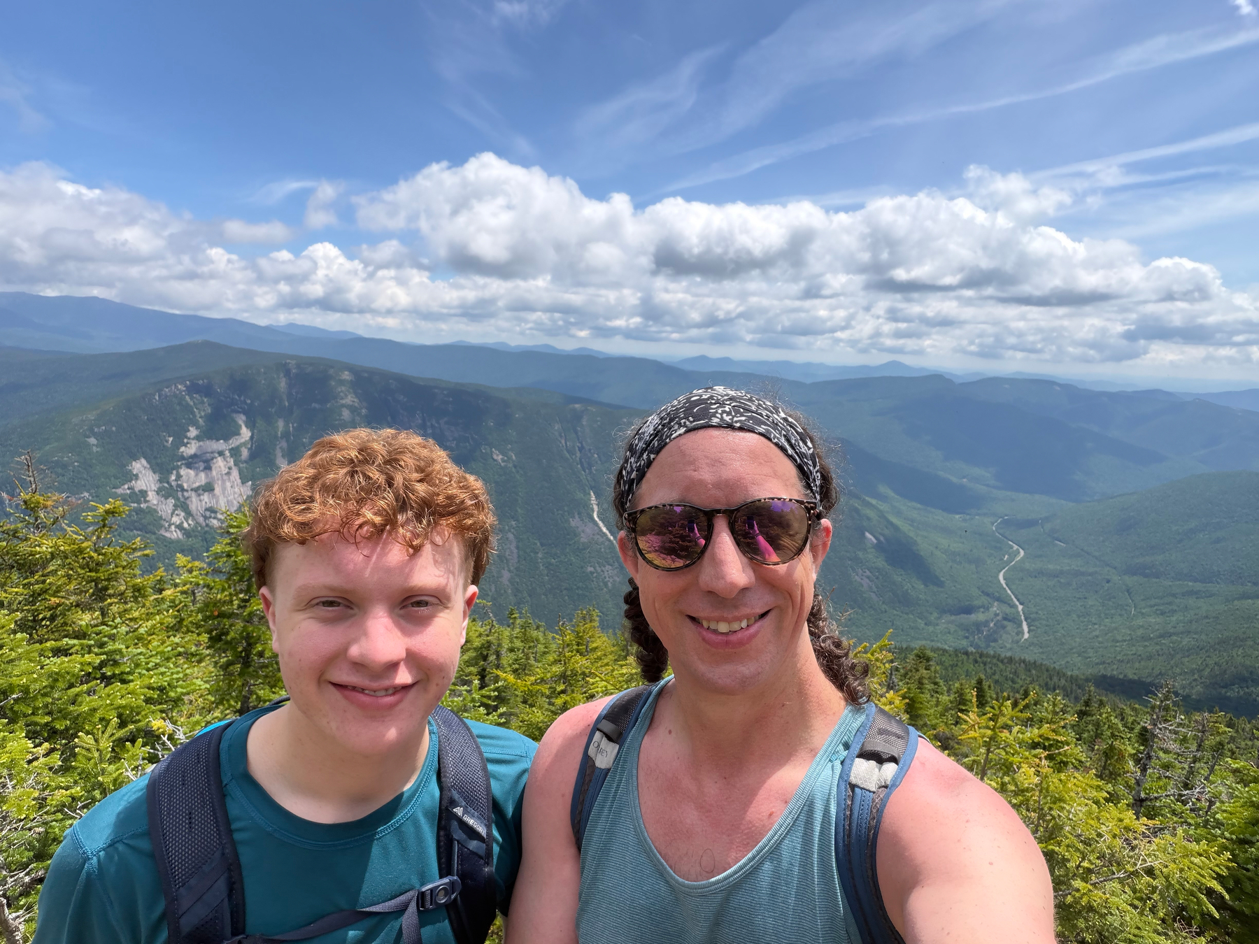 Two people wearing backpacks are smiling at the camera with a scenic mountain landscape and a partly cloudy sky in the background.