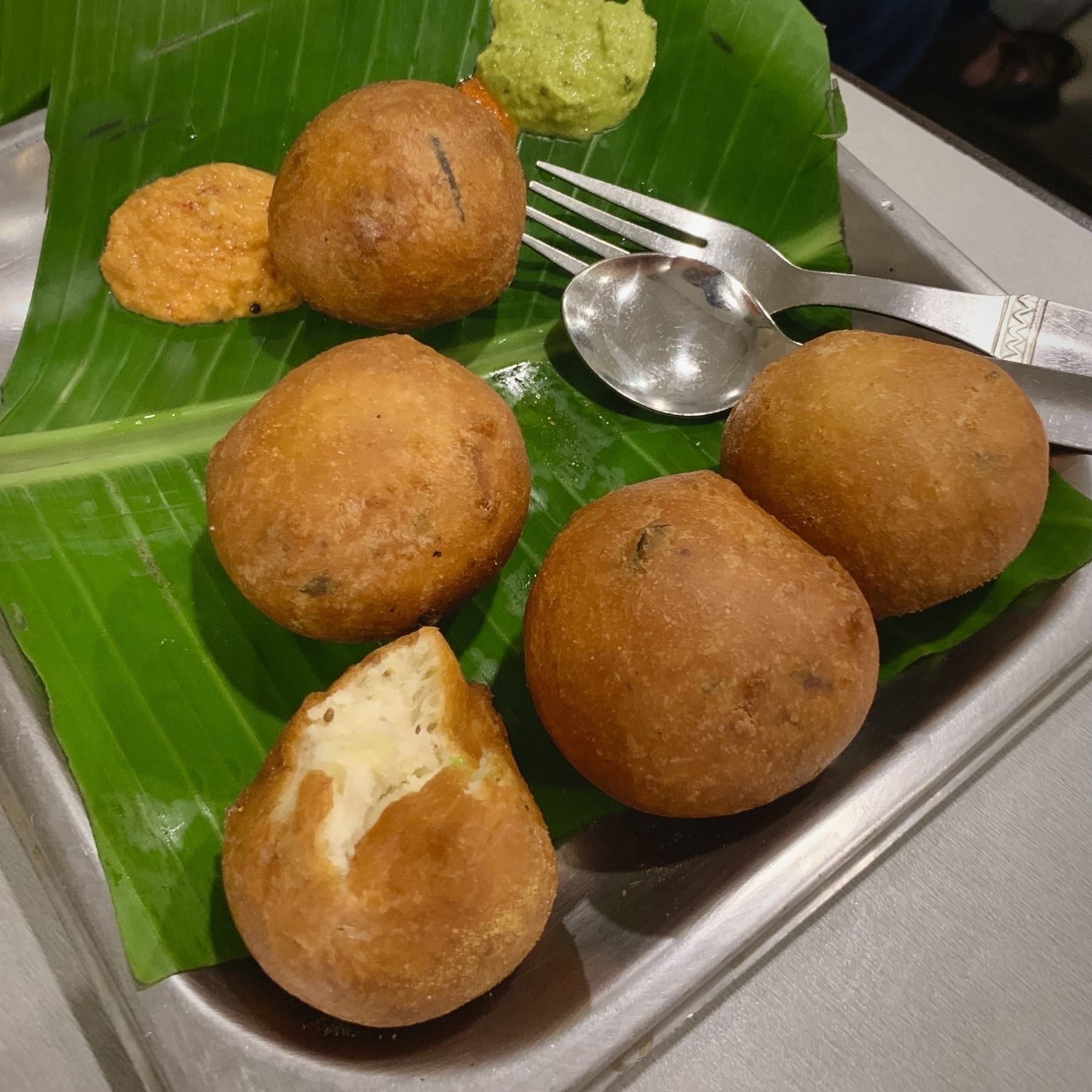 Four Mysore bondas are placed on a banana leaf with two types of chutney, accompanied by a fork and spoon.