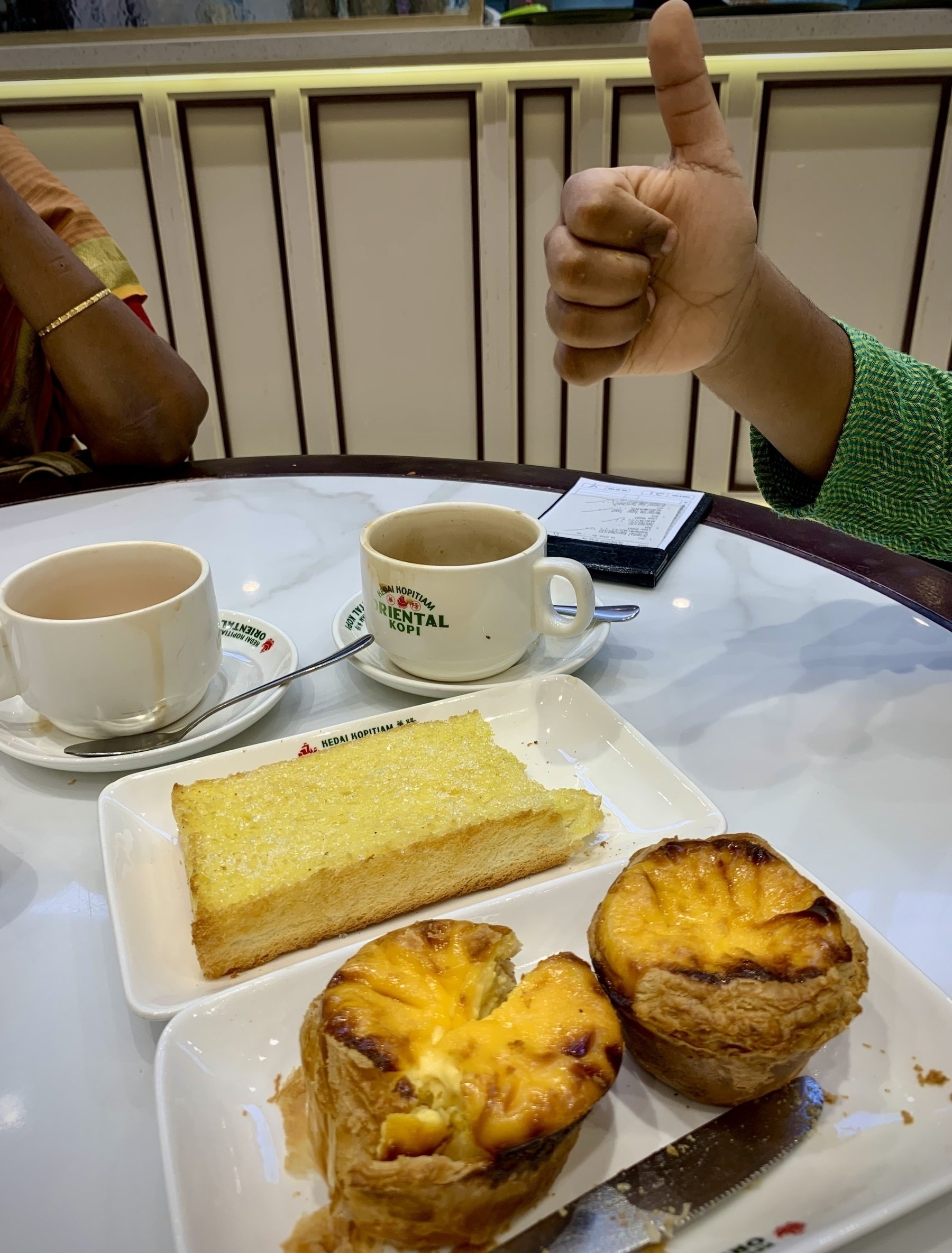 Two cups of coffee, a slice of sugar sprinkled butter toast, and two egg custard tarts are on a table with a boy giving a thumbs-up in the background.