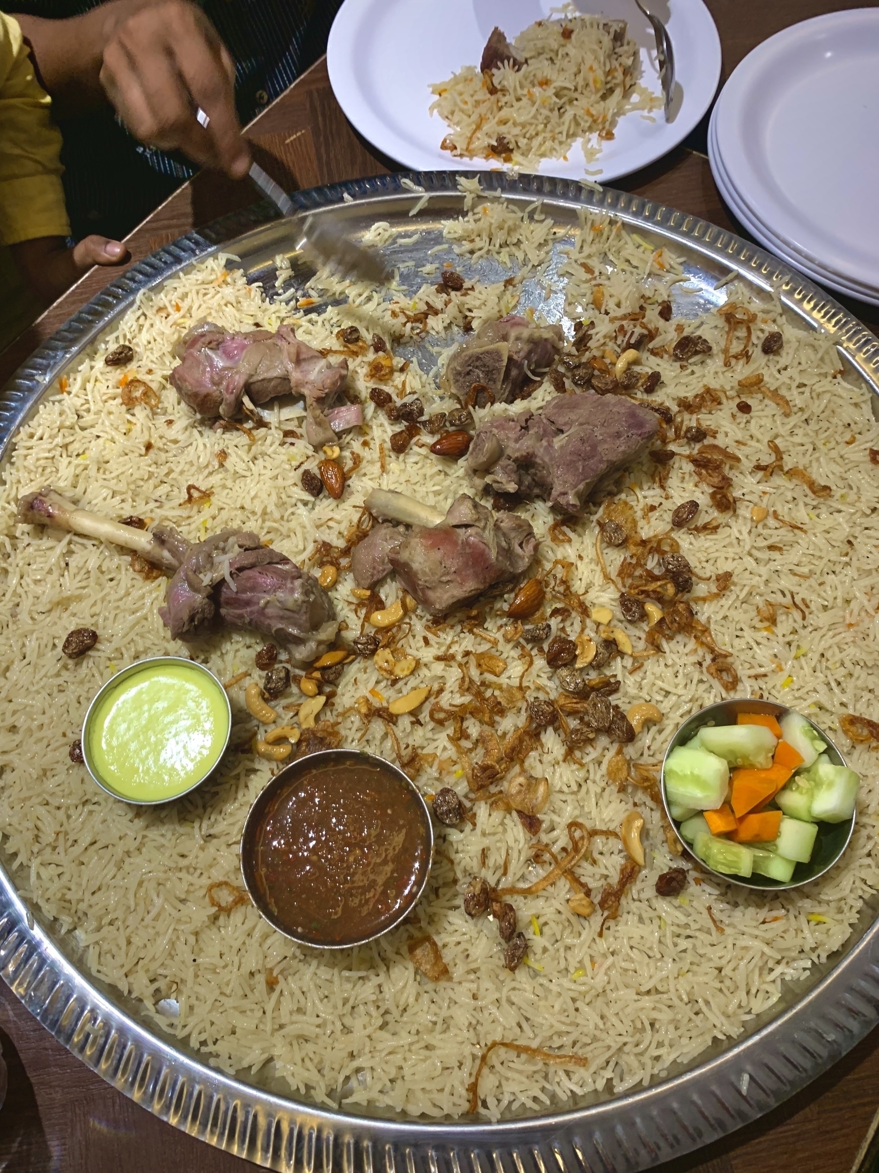 A large platter of Mandi and meat is surrounded by small bowls of sauces and a mix of vegetables.