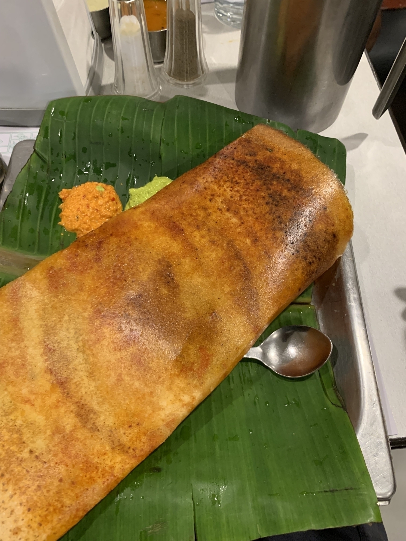 A large, crispy dosa is served on a banana leaf with two types of chutney and a spoon.