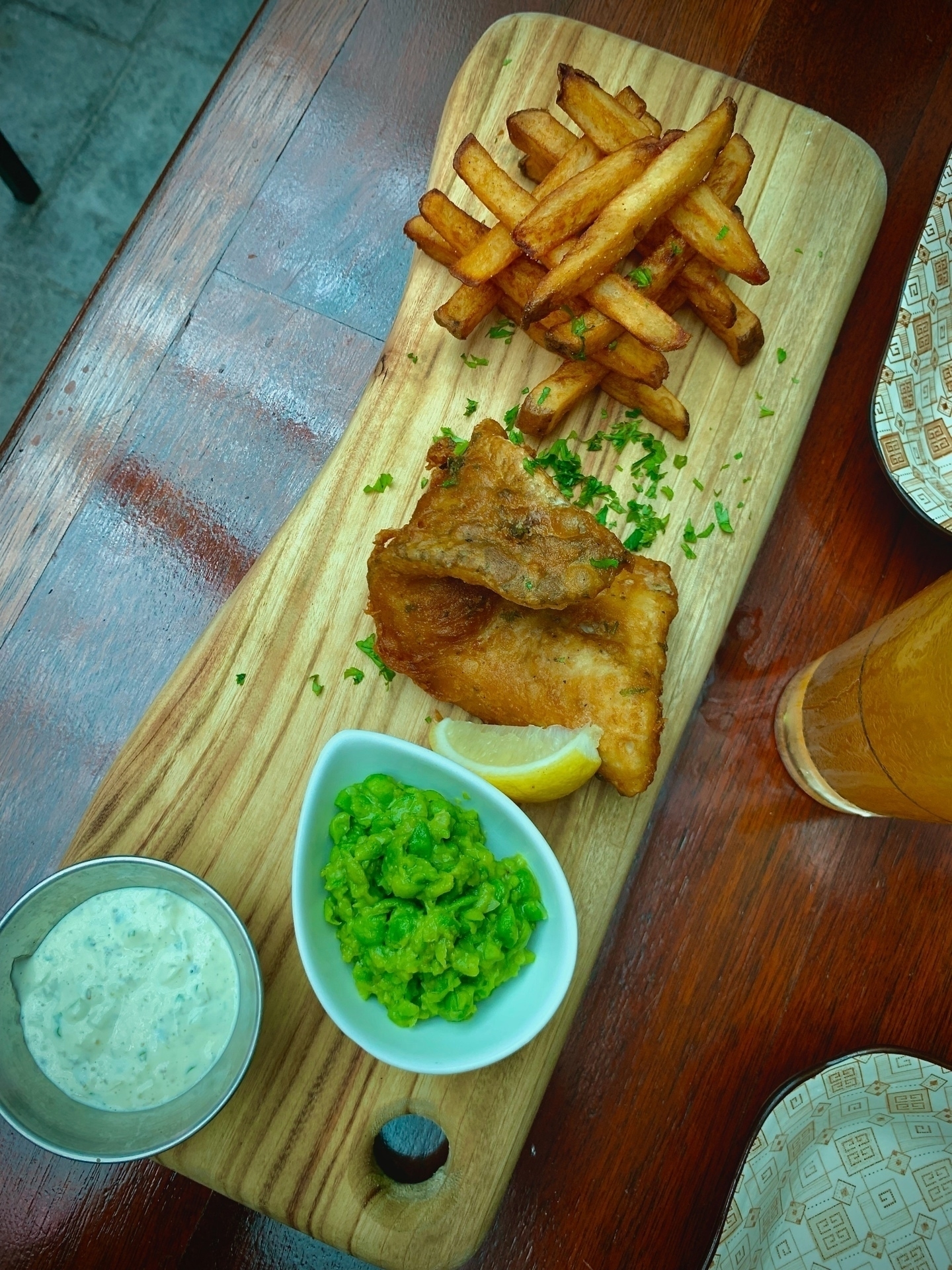 Fish and Chips served on a wooden board, along with mushed peas, a wedge of lemon and tartar sauce