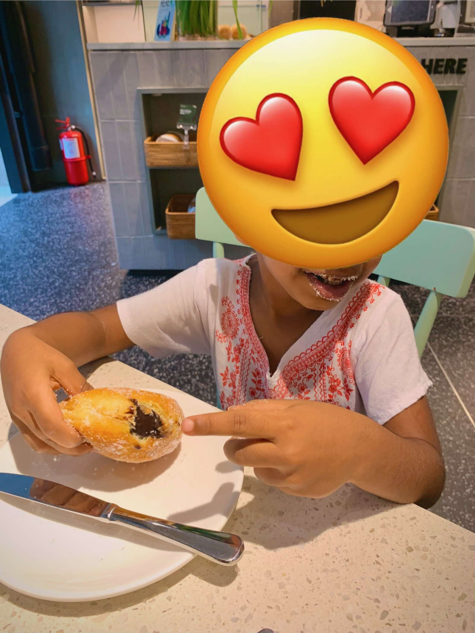 A young boy, whose face masked is with an emoji, enjoying a chocolate filled donut.