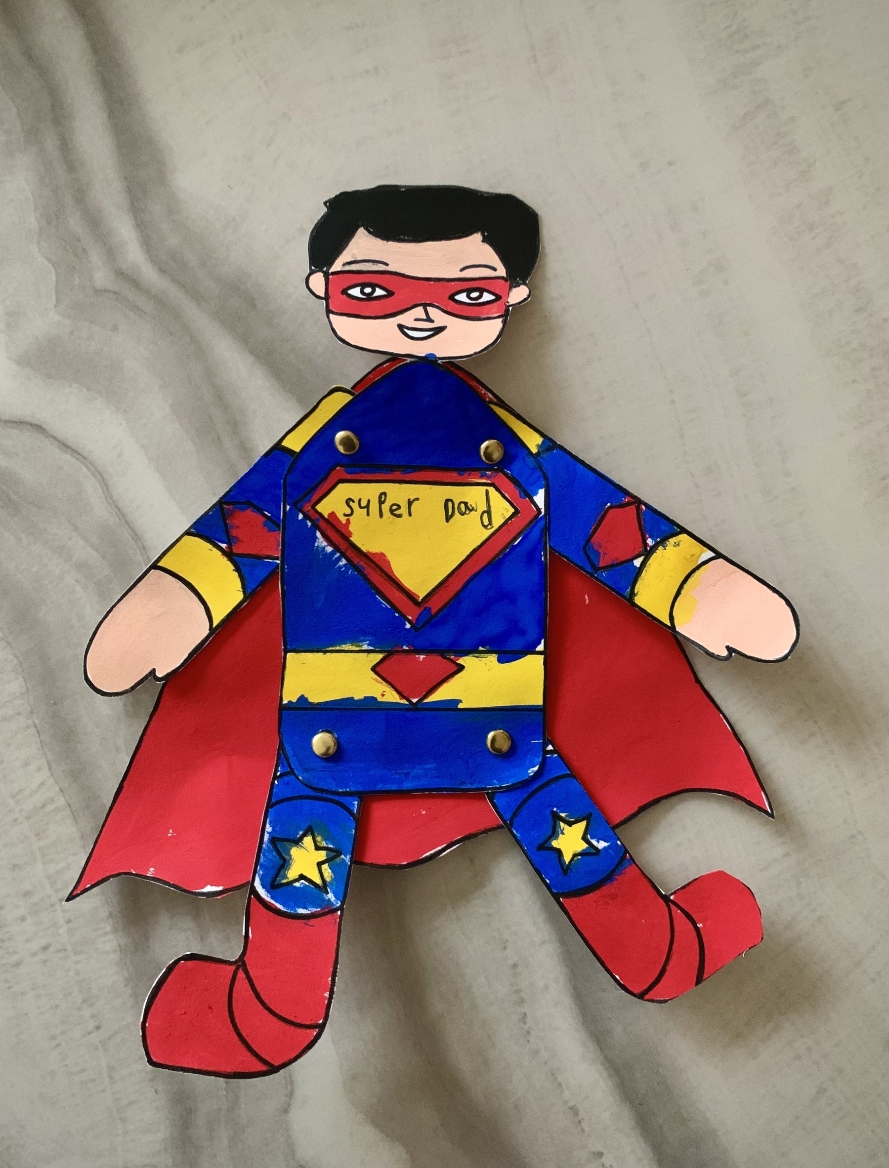 A hand-drawn, colored paper cutout of a superhero with text “Super Dad” on its chest lies on a textured grey background.
