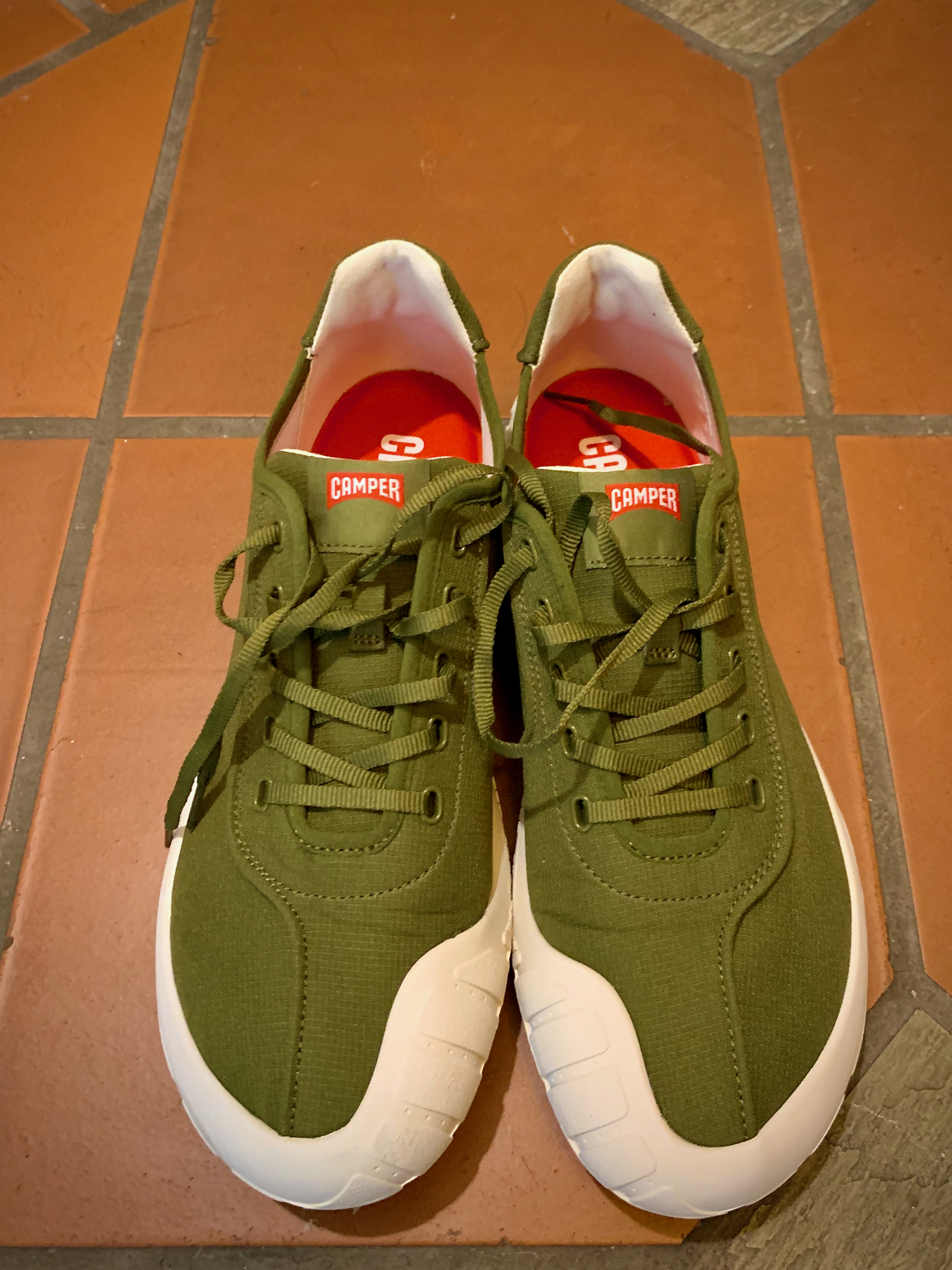A pair of green sneakers with white soles from a brand named Camper, placed on red floor tiles