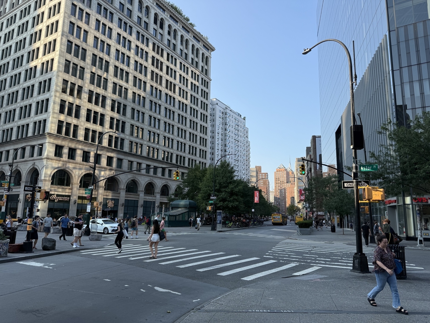 A city intersection in The East Vollage of NYC features pedestrians crossing the street, surrounded by historic and modern buildings under a clear sky.