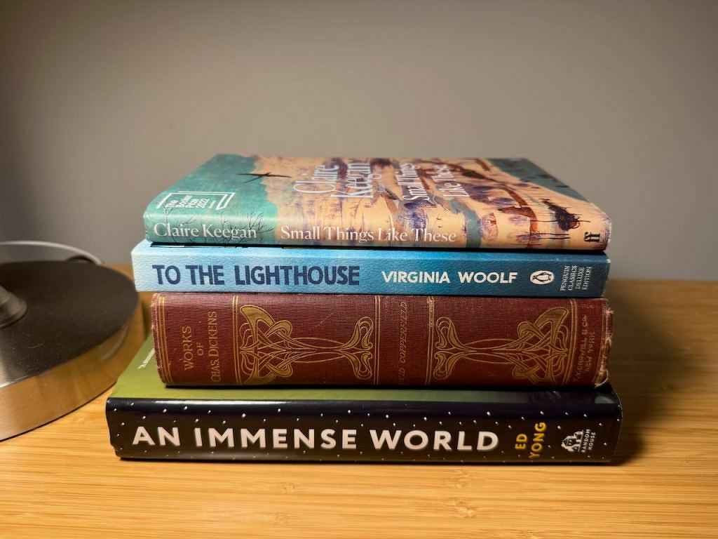 Auto-generated description: A stack of four books, including titles by Claire Keegan, Virginia Woolf, and Ed Yong, is placed next to a lamp on a wooden surface.