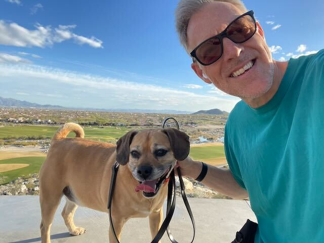 A smiling man wearing sunglasses poses for a selfie with a happy dog on a leash against a scenic outdoor backdrop.