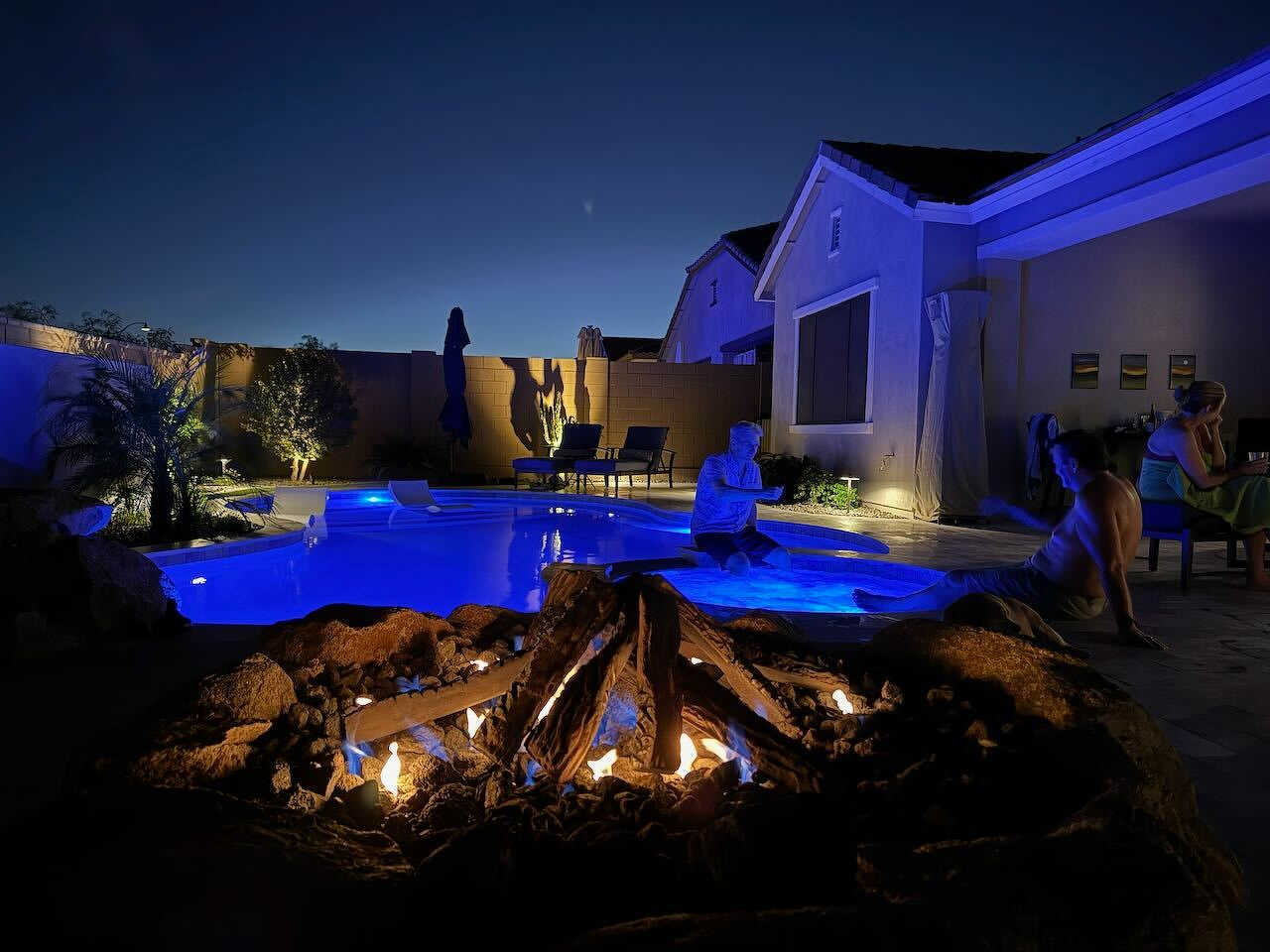 A backyard scene at night features a glowing fire pit in the foreground withA backyard scene at night features a glowing fire pit in the foreground with people relaxing by a brightly lit swimming pool. people relaxing by a brightly lit swimming pool.