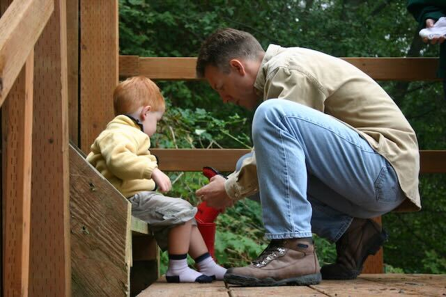 Auto-generated description: A man is kneeling and putting a red sock on a young boy who is sitting on wooden stairs outdoors.