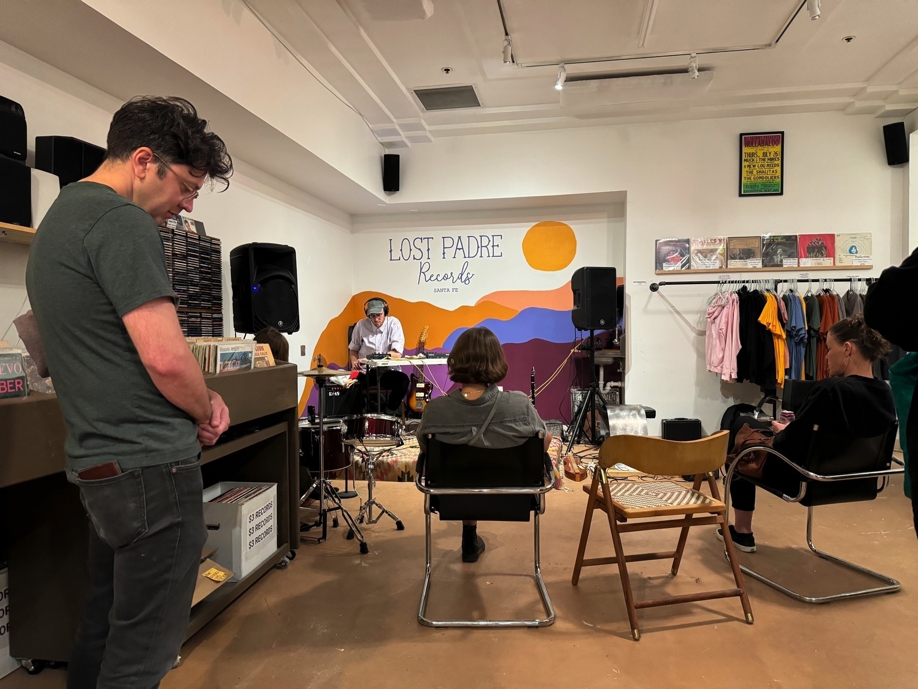 Man seated at table playing field recordings and noise on stage against Lost Padre Records, Santa Fe mural with seated and standing audience members between record store shelves