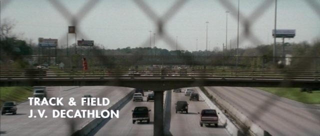 Still from the film Rushmore picturing Max Fisher in a red track suit running across a freeway overpass with the title ‘Track & Field J.V. Decathon’ in the bottom left corner