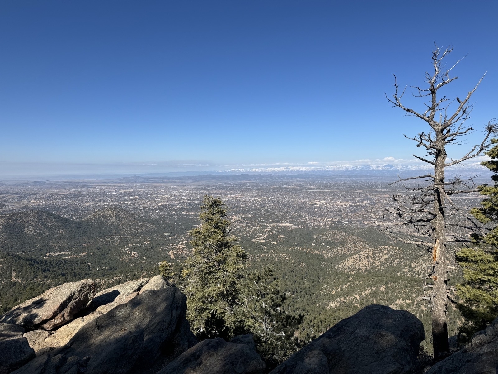 View from mountain peak with rocks and trees in foreground and city of Santa Fe below with snow-covered Jemez mountain range in distant background