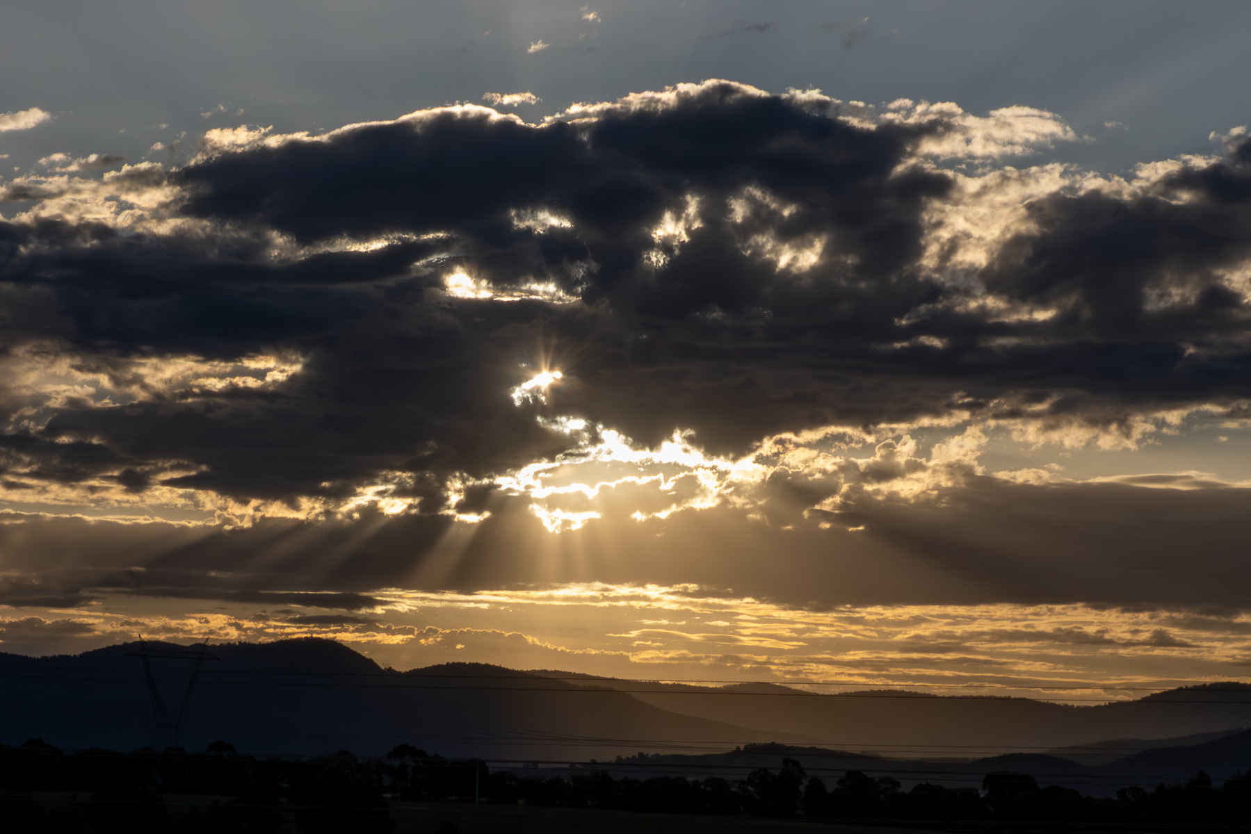 A cloudy sunset with sun rays peaking through the clouds. There are the silhouettes of mountains at the bottom of the image and a sliver of pale blue sky at the top.