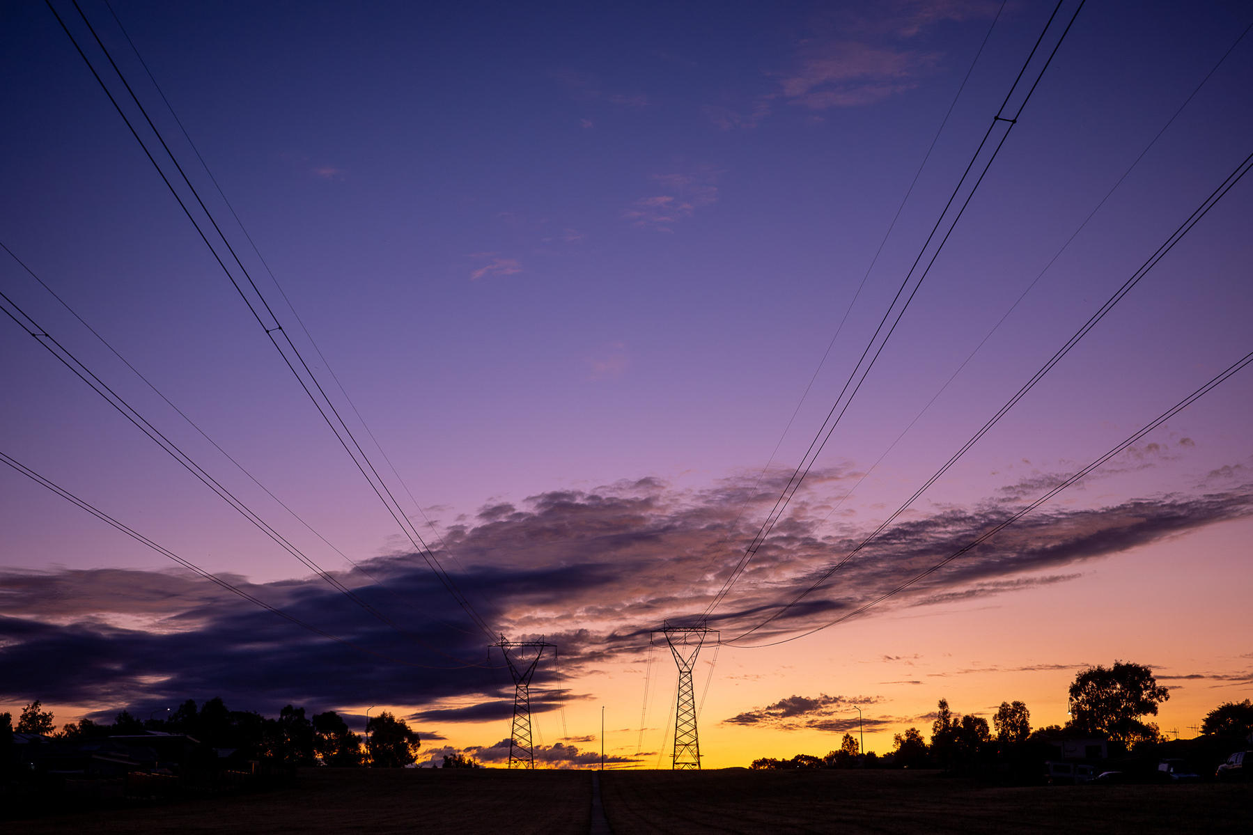 The last of the sunset. A strong orange and yellow glow on the horizon, sark grey clouds, purple and deep blue sky. There are high voltage powerlines as leading lines from the top corners of the image to the centre.