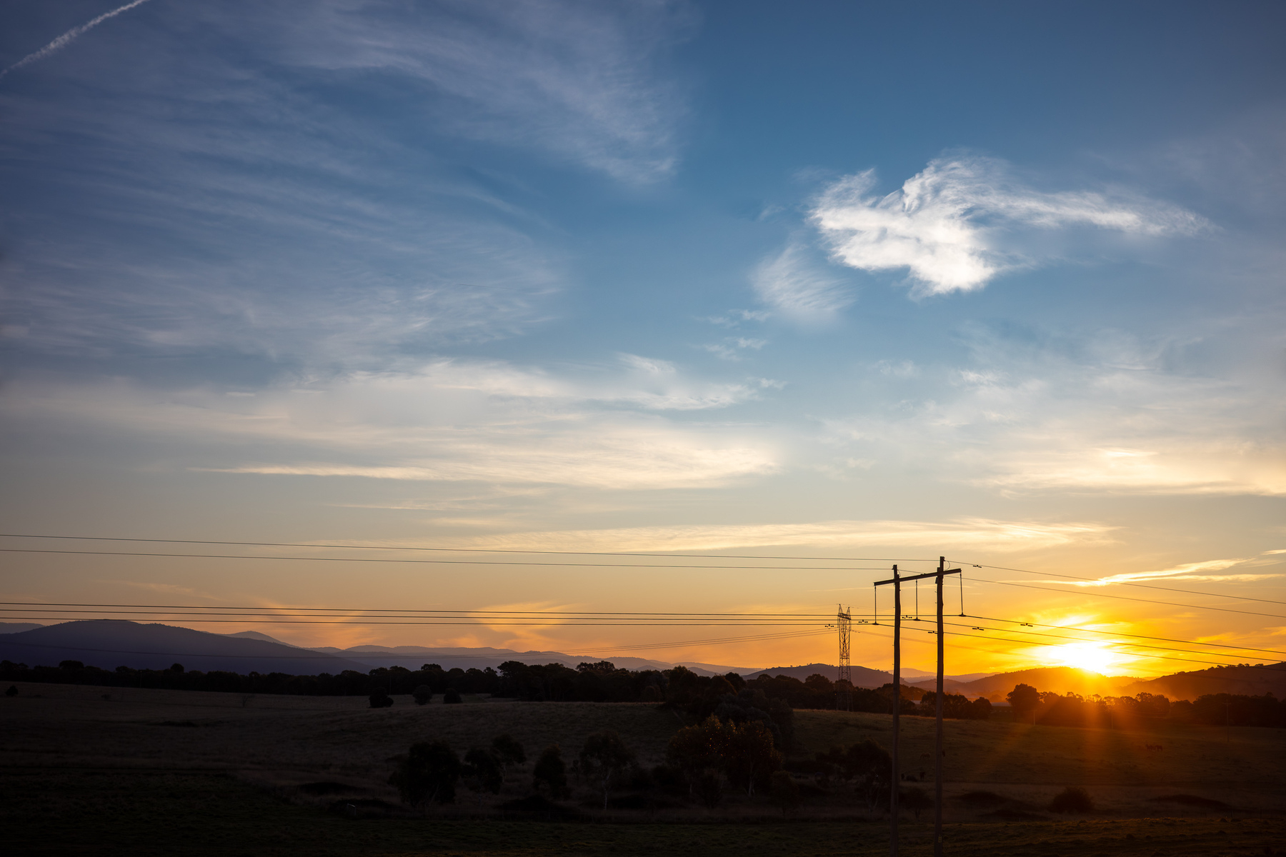 Sunset. Yellow and orange sun setting with a blue sky with wispy clouds and powerlines on the horizon.