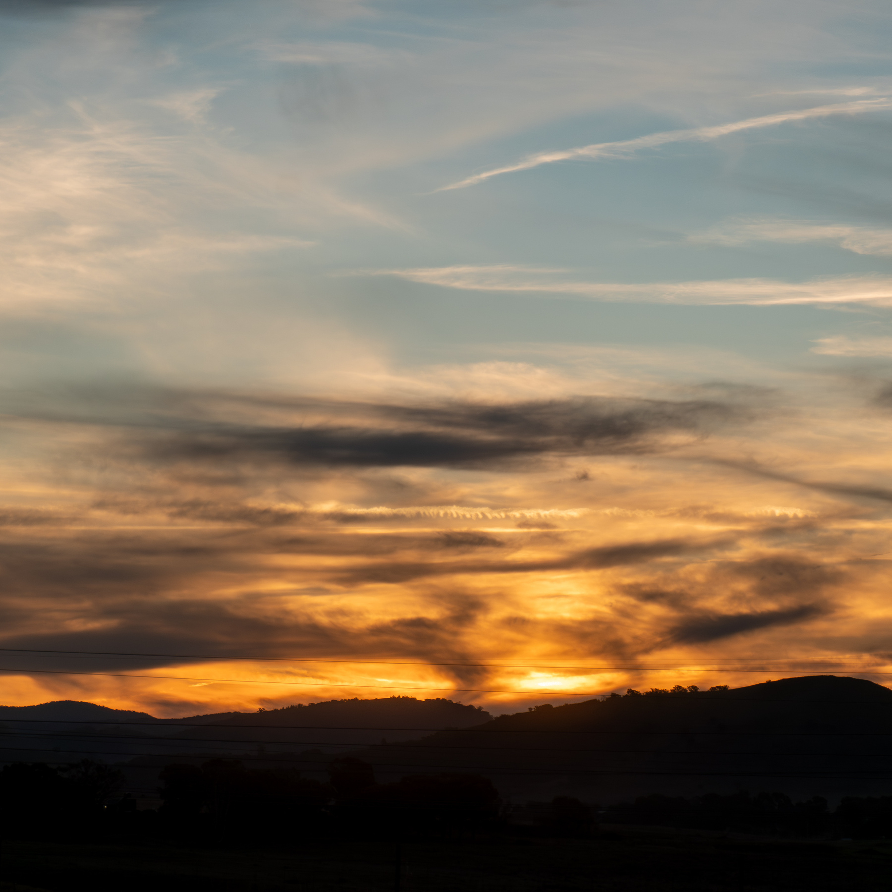 Sunset. A yellow and orange glow above the silhouette of the hills on the horizon. Grey and white clouds above the horizon with a light blue sky.