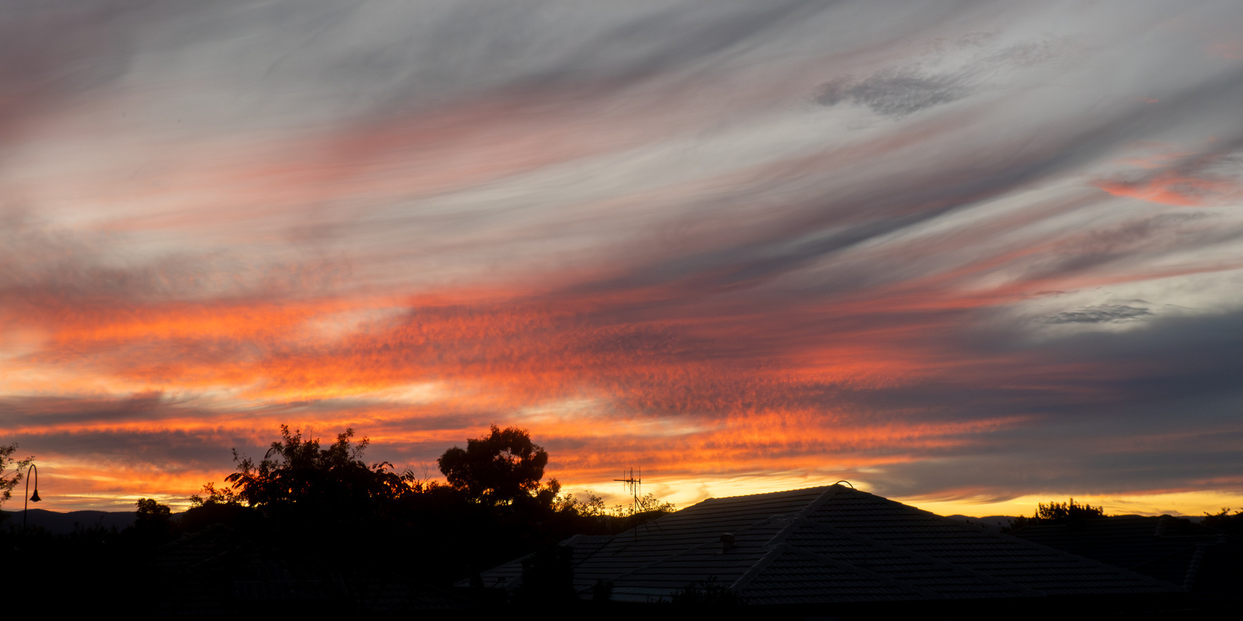 Sunset - silhouette of houses and trees at the bottom - orange and yellow glow with streaky grey clouds.