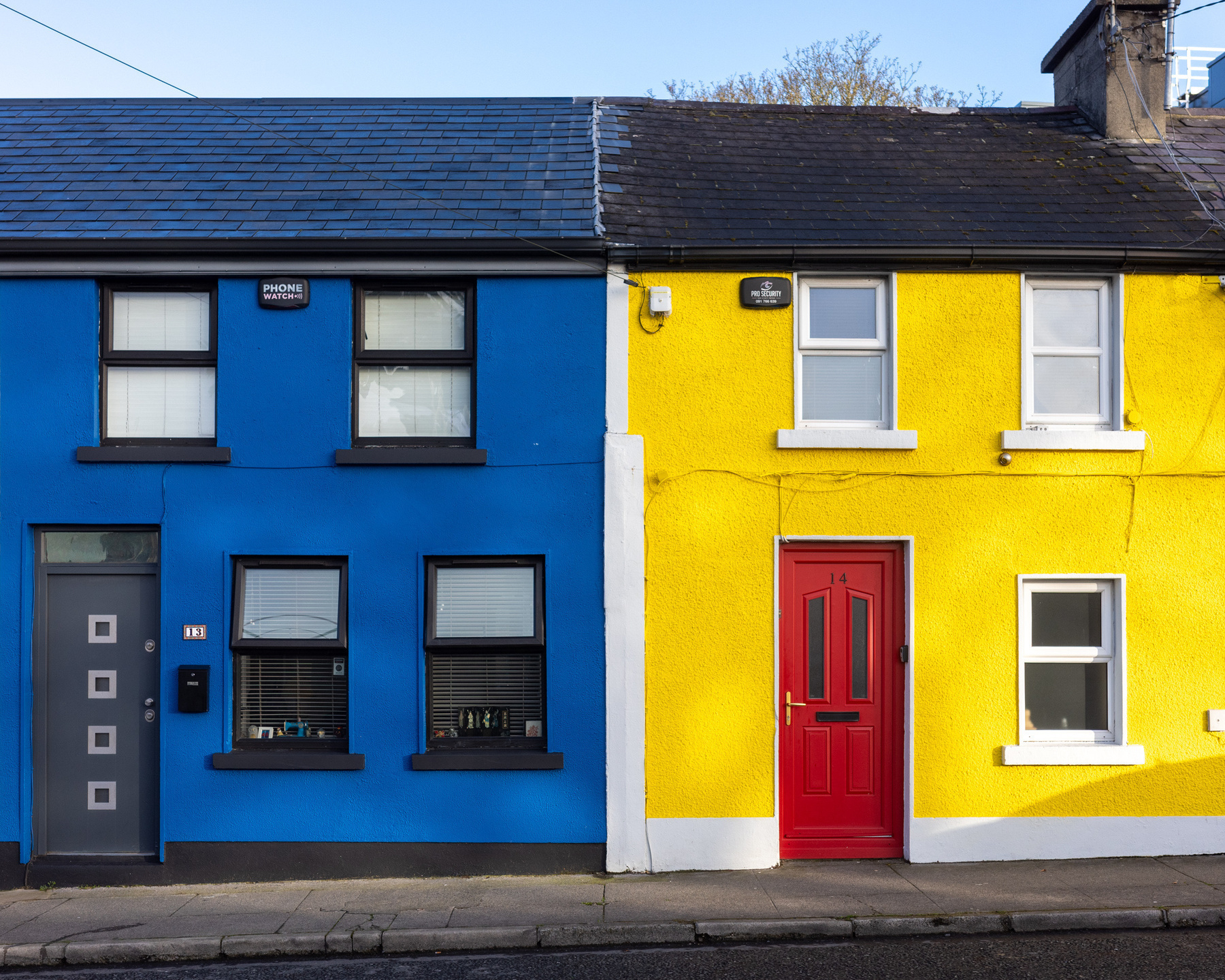 A pair of terrace houses. The one of the left is painted blue, the one on the right is painted yellow and has a red door. They are two storeys tall with two windows on the top storey.