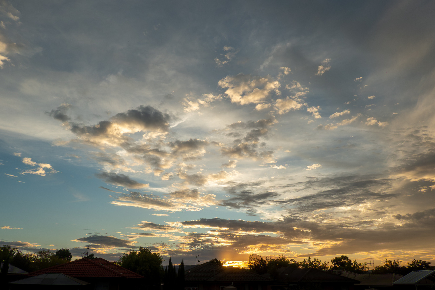 A sunset. Light blue sky. Soft yellow glow from the horizon above the silhouette of house roofs and trees. There are light grey and white clouds throughout the sky.